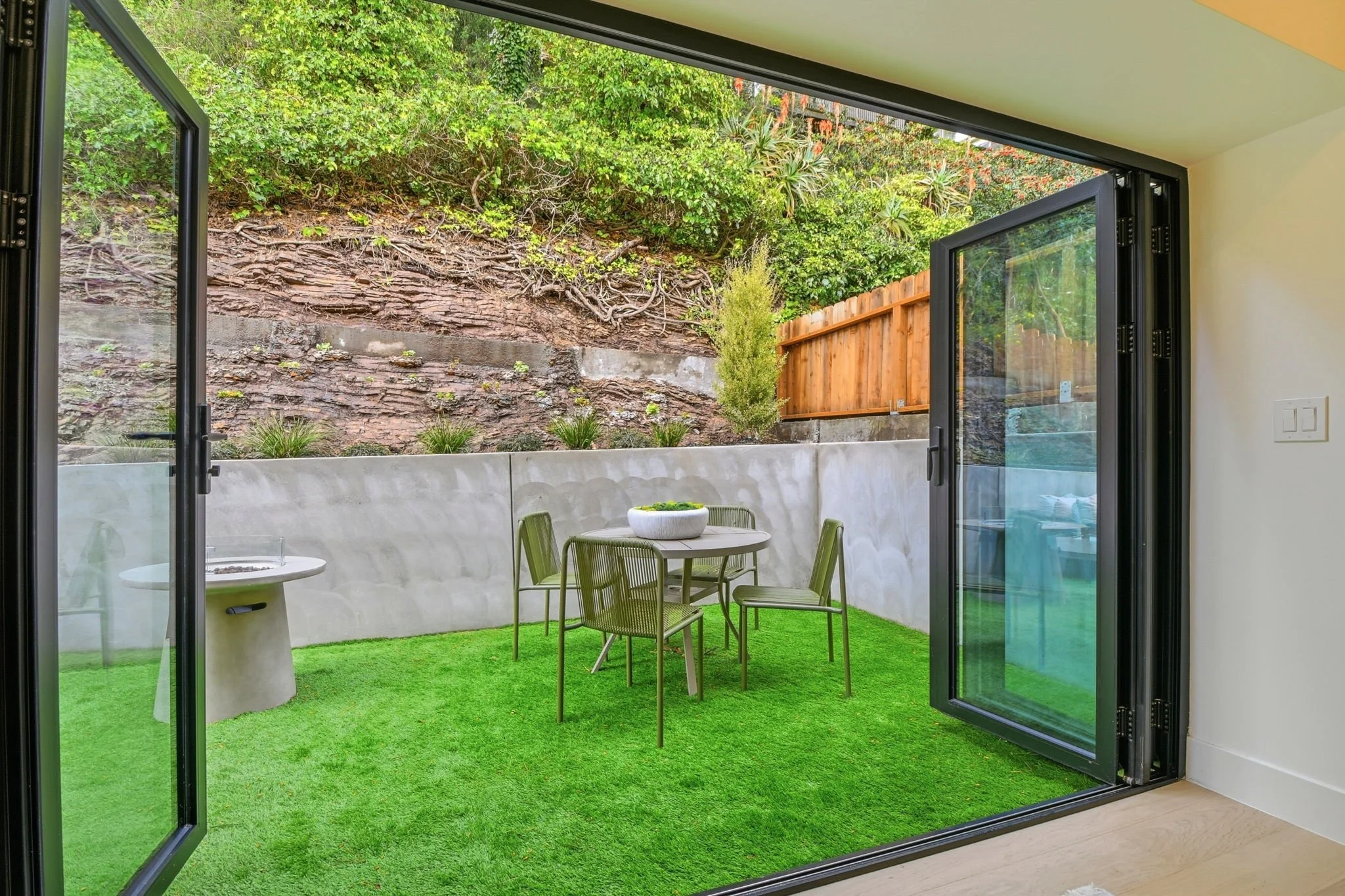 Secluded San Francisco patio area with a fire pit, concrete retaining wall, and lush hillside views framed by massive glass folding doors.