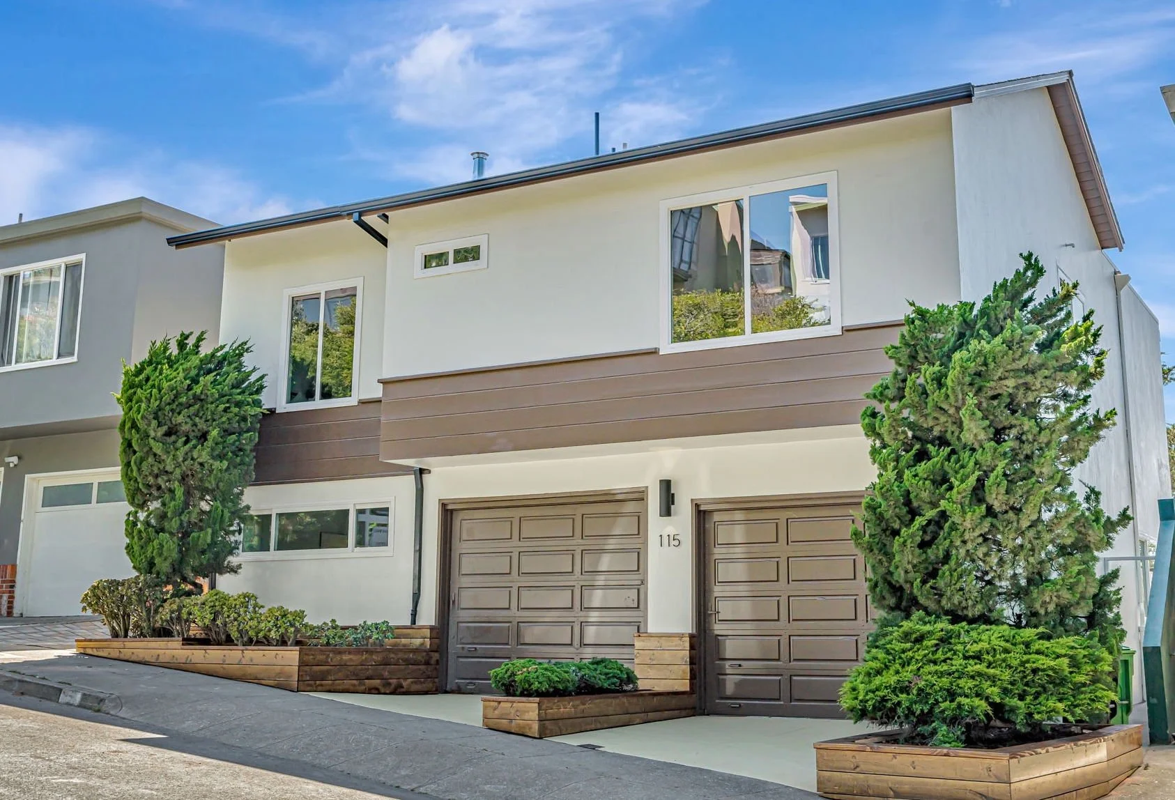 The front exterior of 115 Gladeview Way, a contemporary two-story San Francisco home. The house features white stucco, brown horizontal siding, and two dark brown garage doors. Tiered wooden planters with manicured shrubs line the driveway under a br