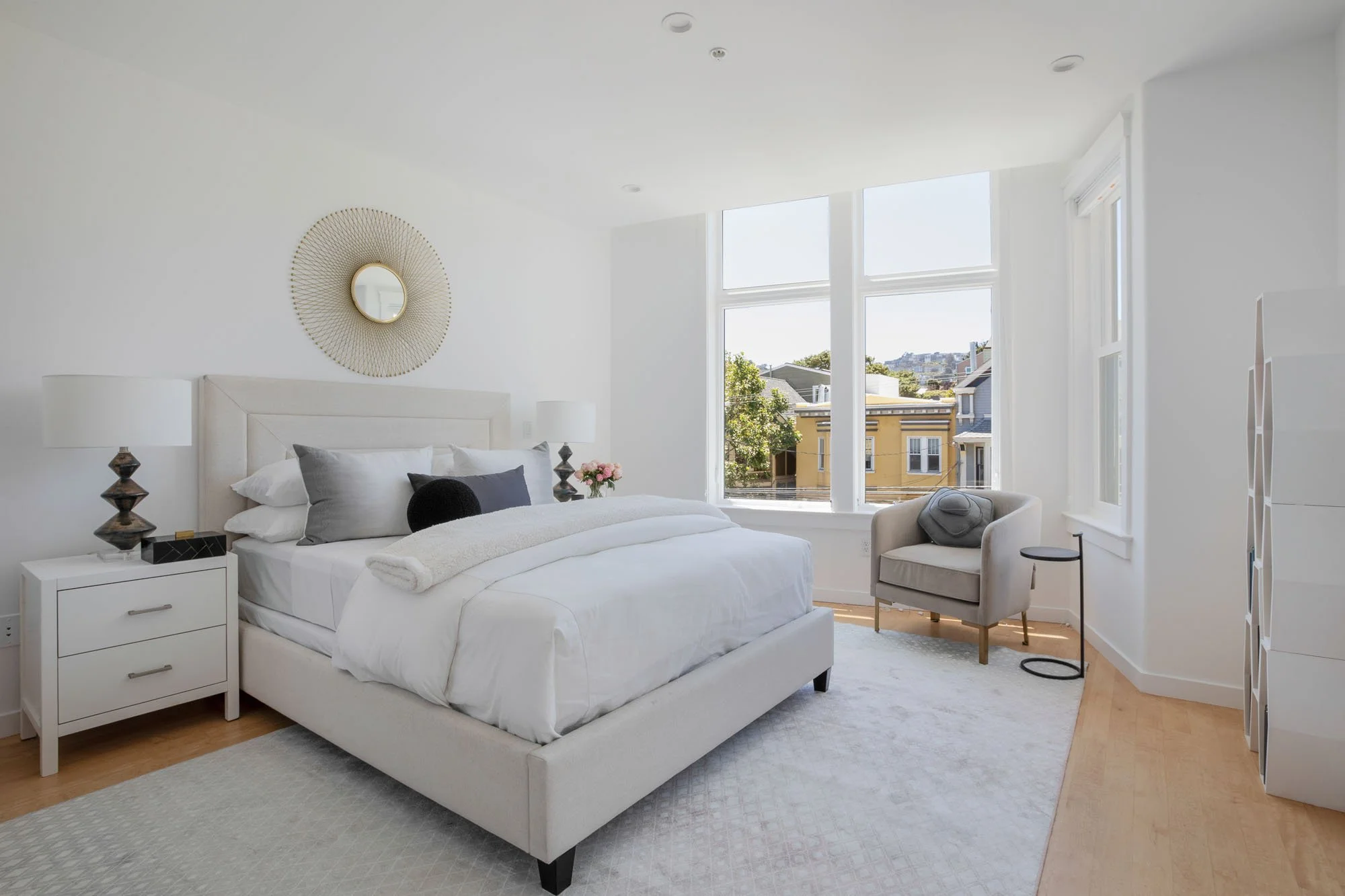 Modern luxury bedroom with a cream upholstered bed, gold sunburst mirror, white nightstands, and large floor-to-ceiling windows overlooking a city neighborhood.