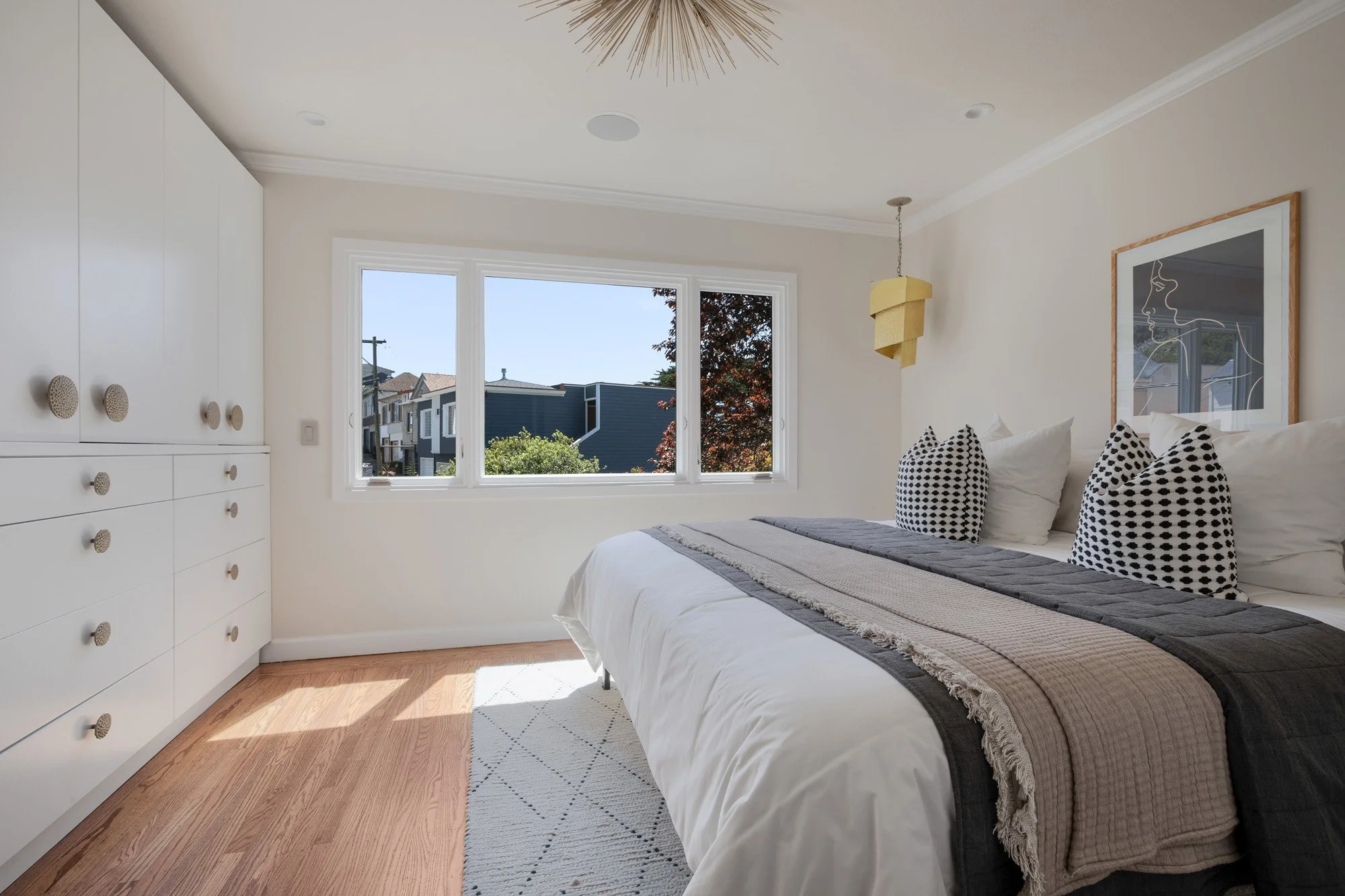 Sun-drenched primary bedroom with large windows, custom white wardrobes, and contemporary gold pendant lighting.