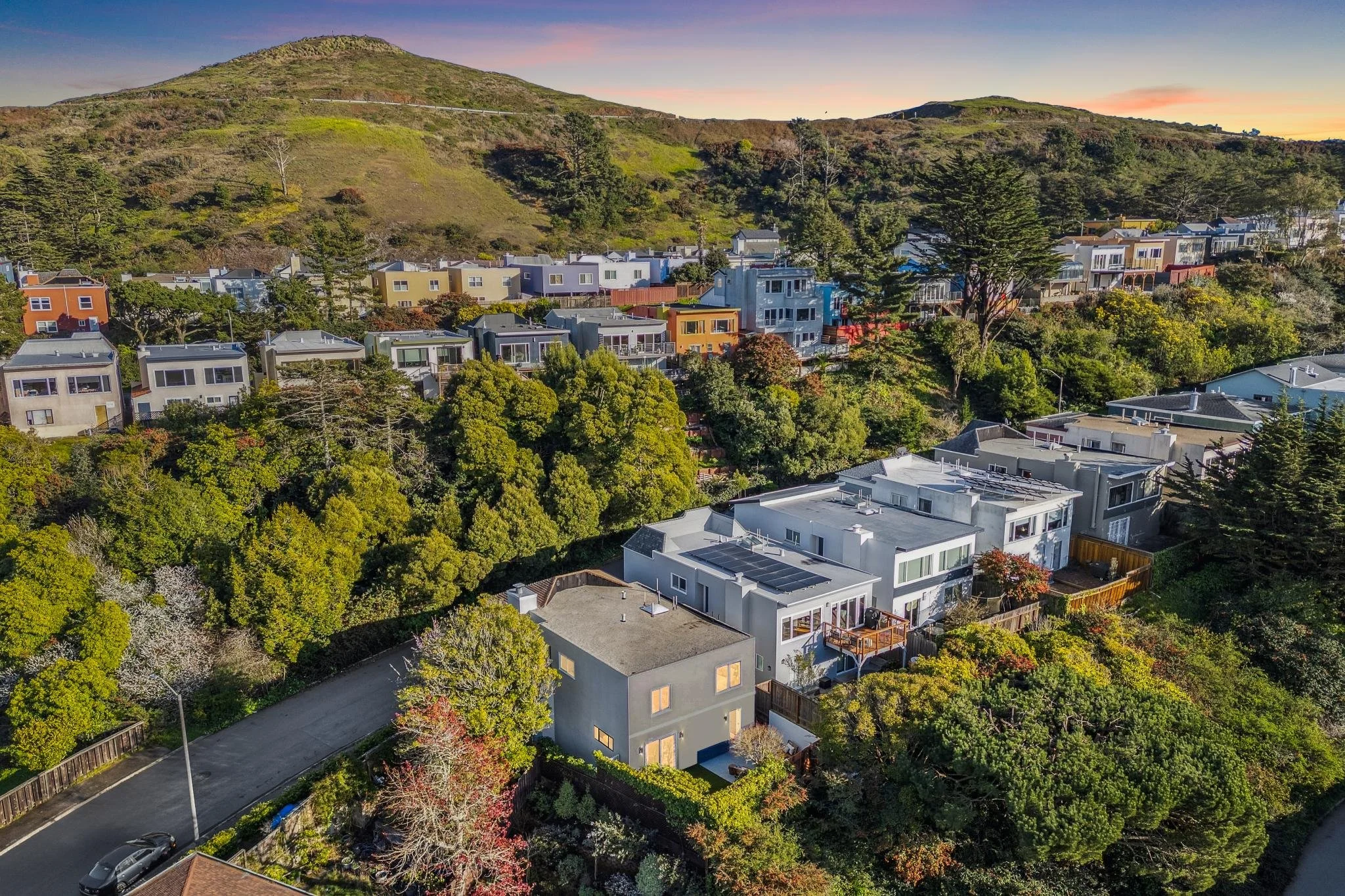 High-angle drone shot of a hillside neighborhood in San Francisco showing a grey modern home with a flat roof and solar panels, surrounded by lush green trees and coastal hills.