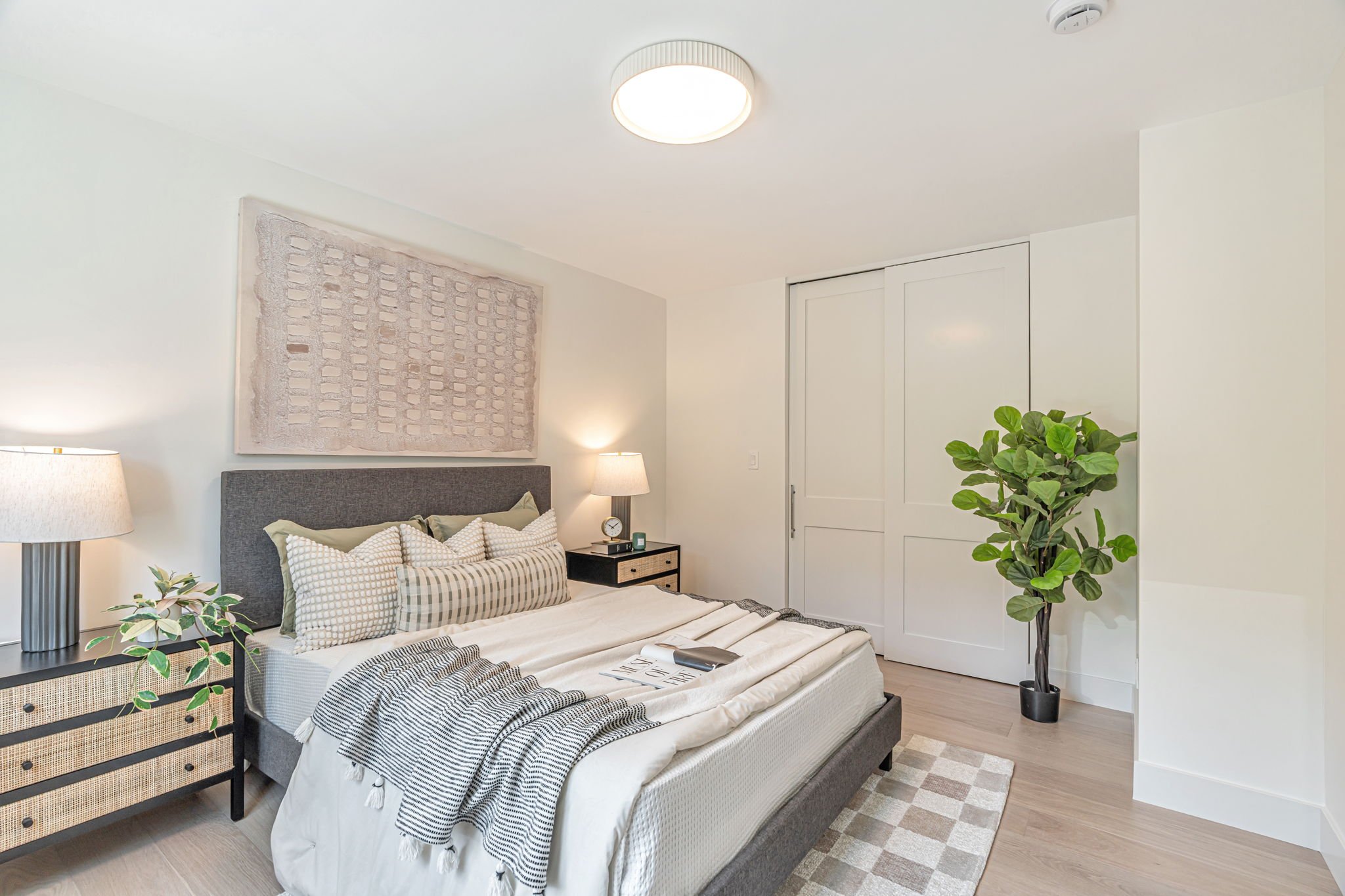 A full view of the primary bedroom featuring neutral walls and light wood floors. The bed is styled with a grey headboard and layered linens, flanked by a tall potted fiddle-leaf fig plant and white sliding closet doors.