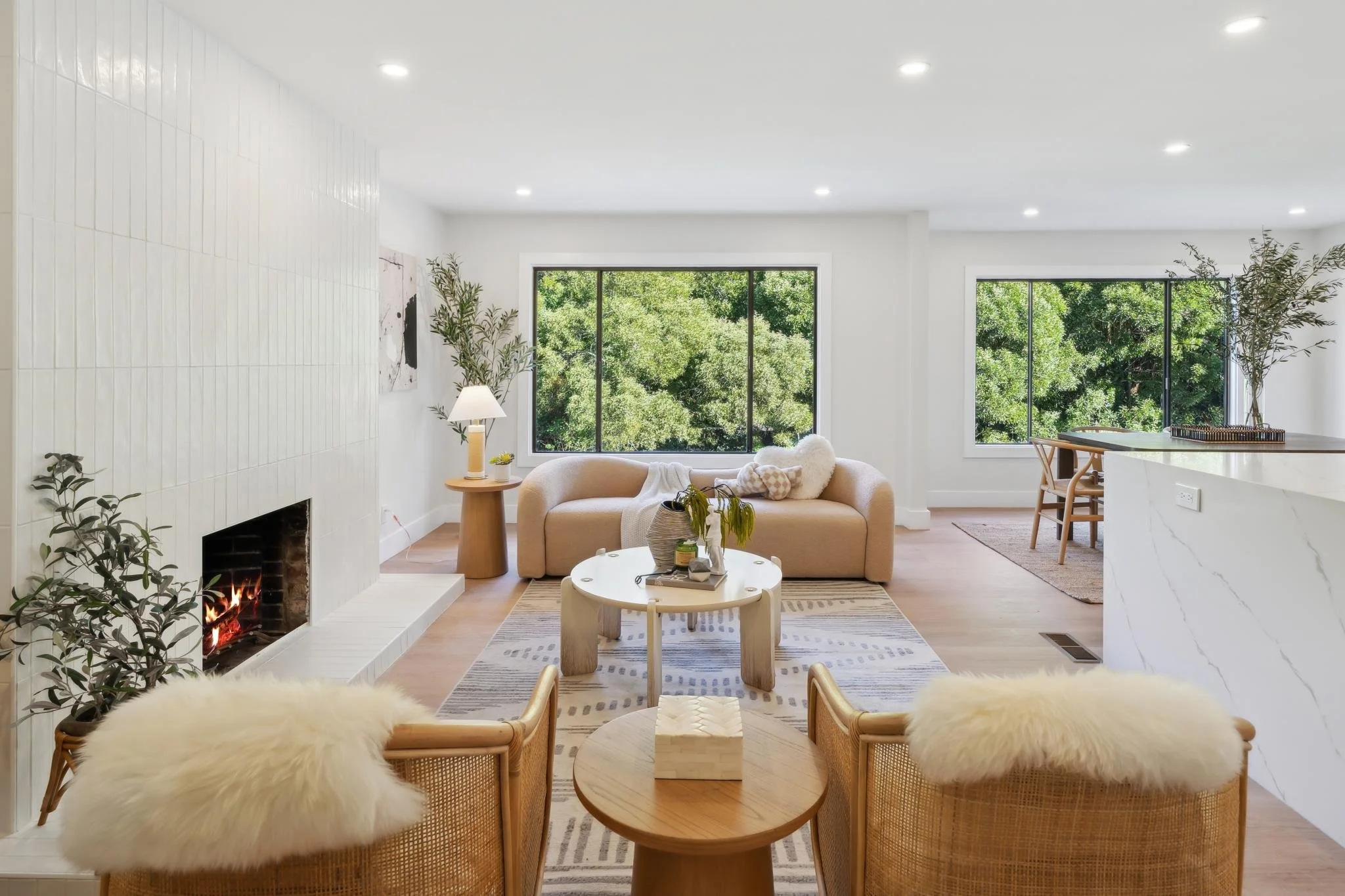 Spacious living room with a white vertical tile fireplace, minimalist furniture, and large black-framed windows offering a lush view of green trees.