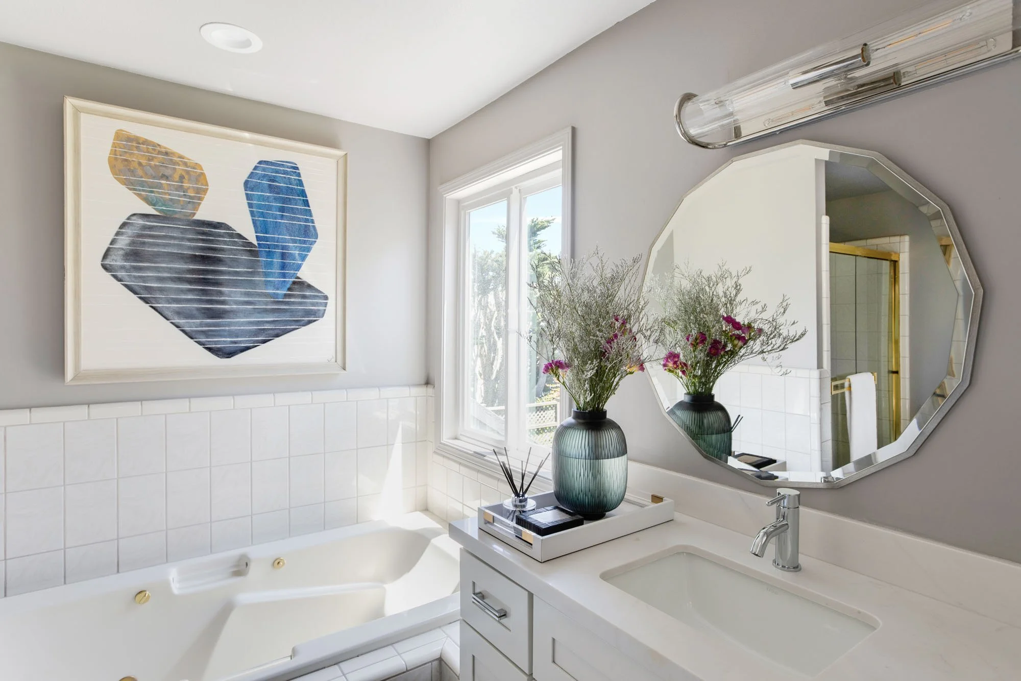 Bright bathroom interior highlighting a modern octagonal mirror, designer light fixtures, and a clean white vanity, with abstract wall art and a view of the outdoor greenery.