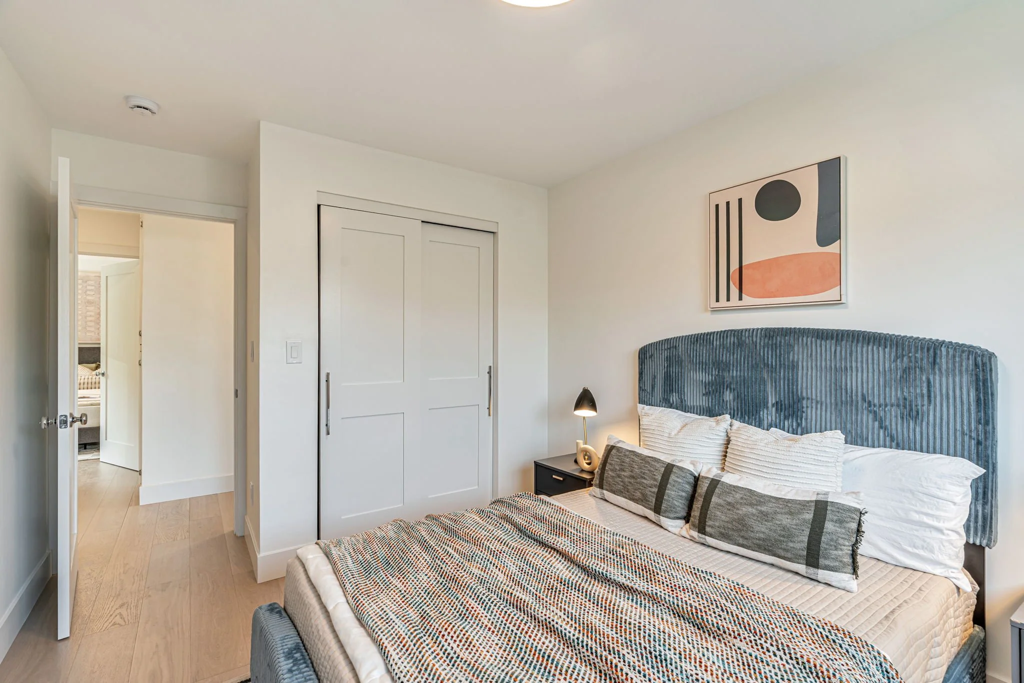 A third bedroom featuring a unique blue corduroy-textured headboard and modern abstract art. The room has a clean white interior with a sliding closet door and a view into the hallway.