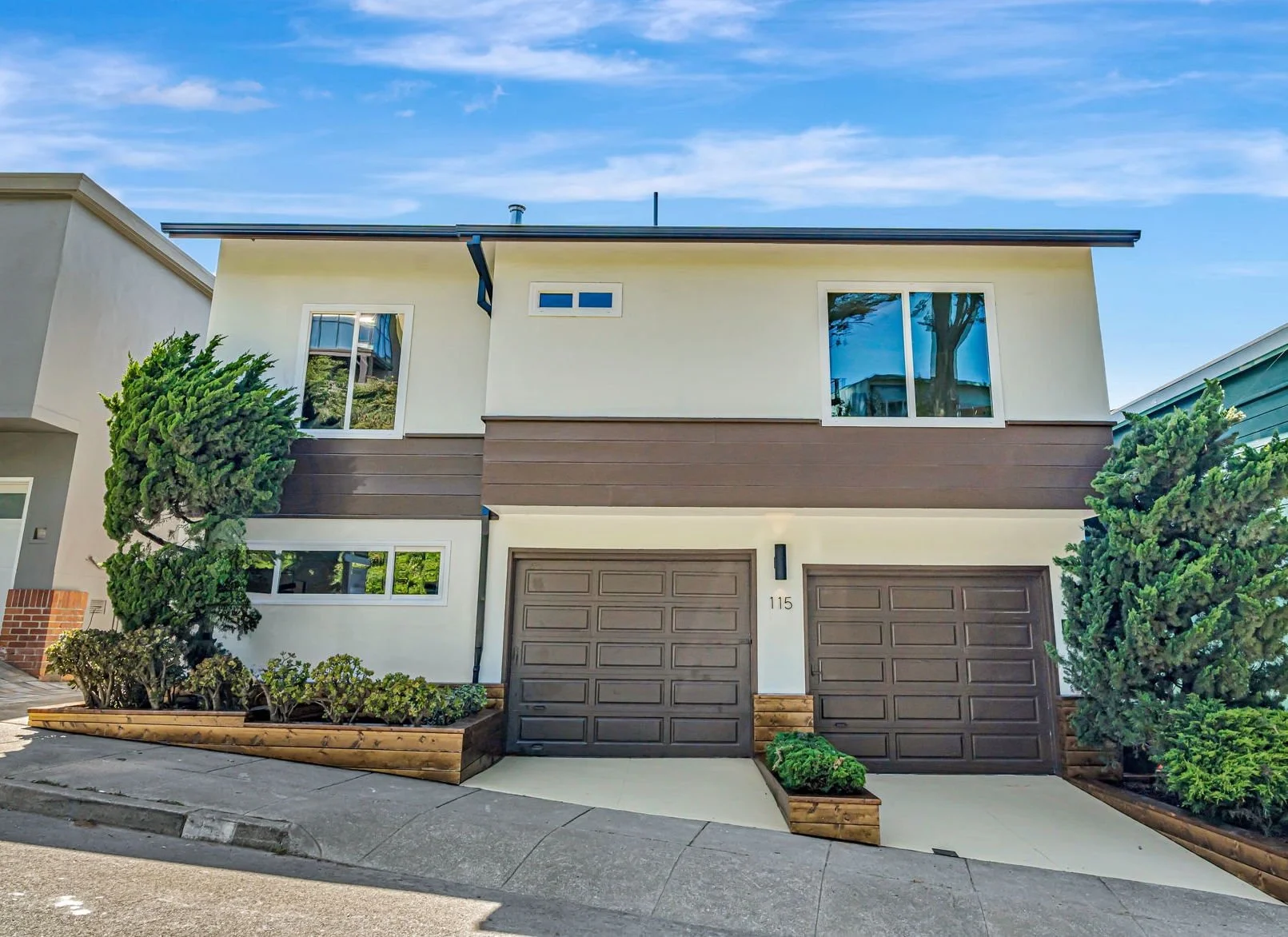 A direct front view of 115 Gladeview Way, a two-story residence showing the symmetrical garage doors and upper-level windows. The modern architecture is accented by dark trim and a clean white facade with lush green trees on either side.