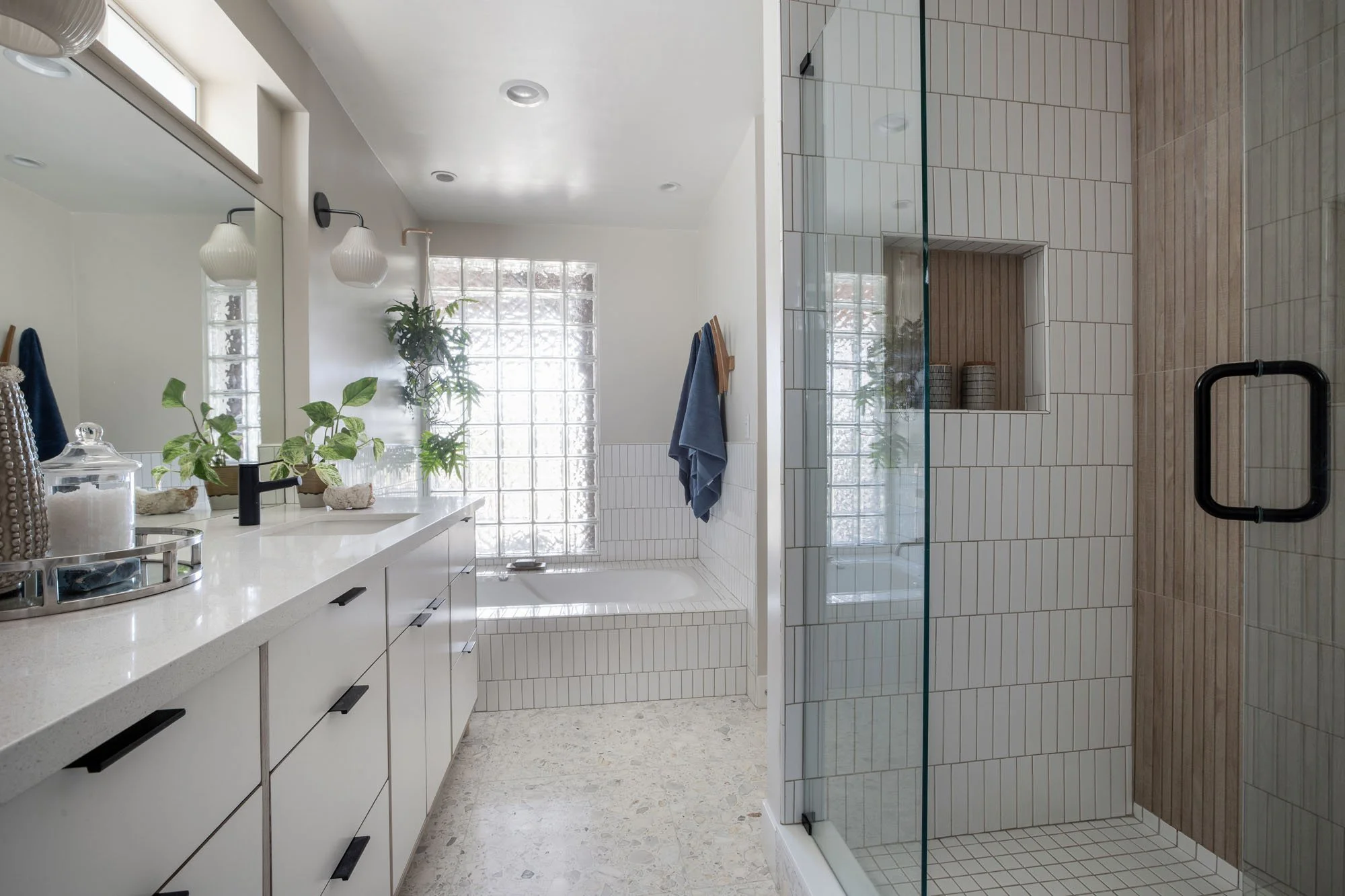 Modern bathroom featuring a white double vanity, glass-enclosed walk-in shower with vertical tile, and a large glass block window over a soaking tub.