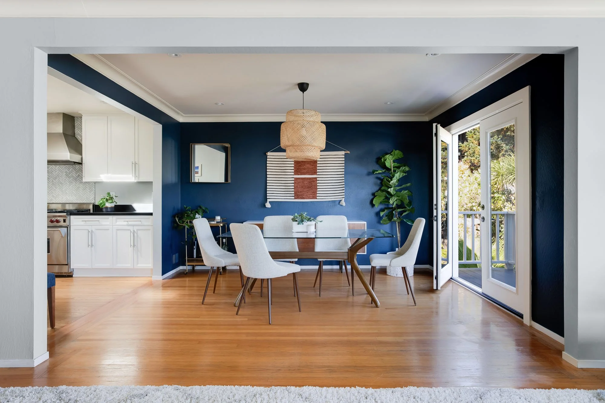 Formal dining room with a navy blue accent wall, a glass-top table with mid-century chairs, a woven pendant light, and French doors leading to a private deck.