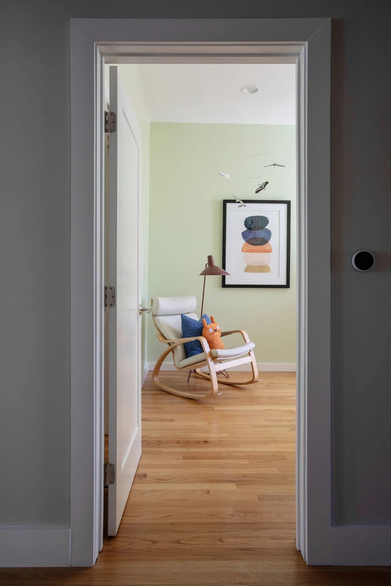 View through an open door to a light green nursery featuring a modern rocking chair, hardwood floors, and abstract wall art.
