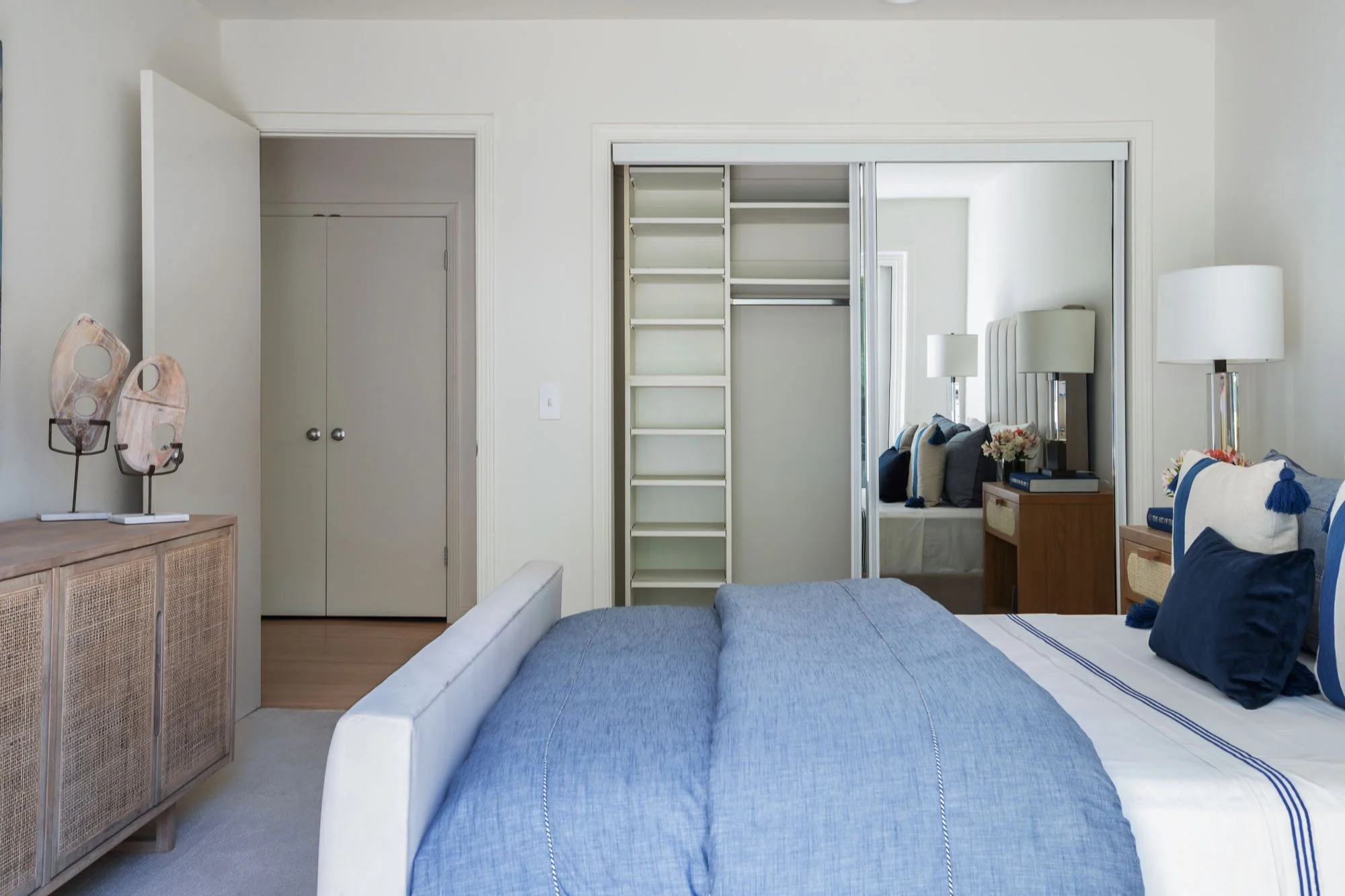 Modern bedroom interior with an open mirrored sliding closet featuring custom built-in shelving, a light wood cane dresser, and high-end coastal decor.