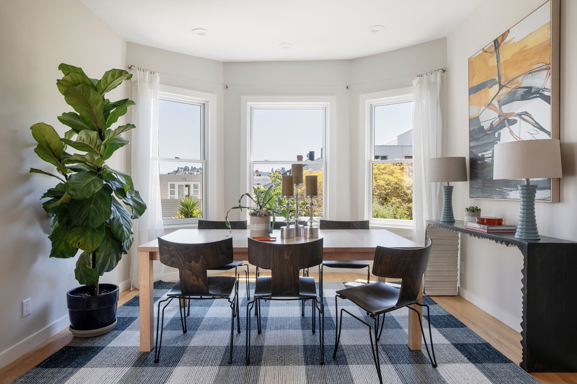 Modern dining room featuring a wood table with black designer chairs, a large fiddle leaf fig plant, and a blue plaid area rug under a bay window.