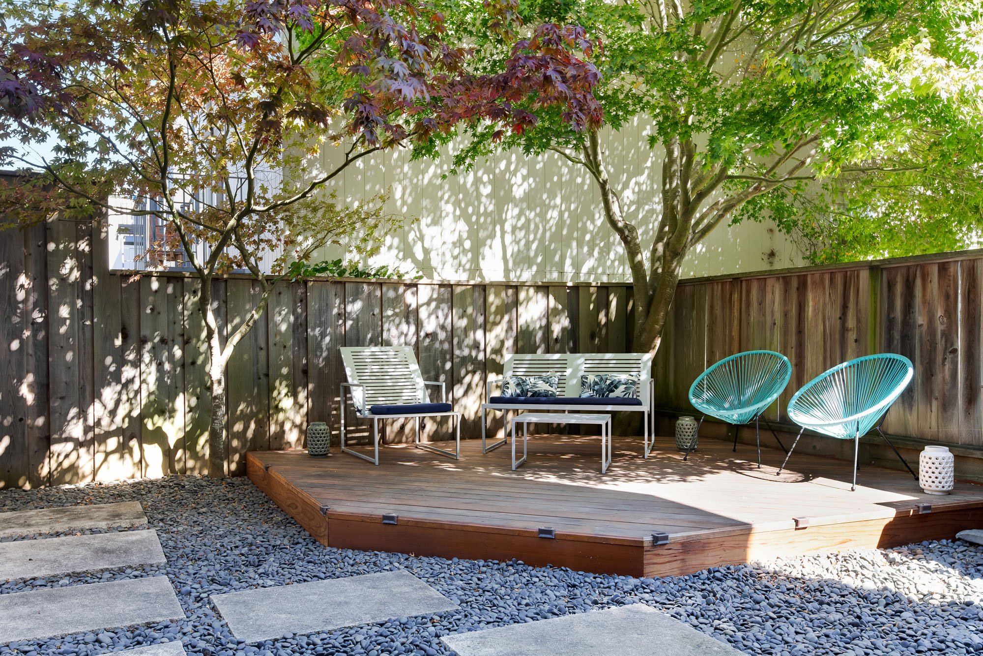 A shared wooden sun deck in a secluded backyard, featuring modern white outdoor furniture, turquoise Acapulco chairs, and a mature Japanese Maple tree.