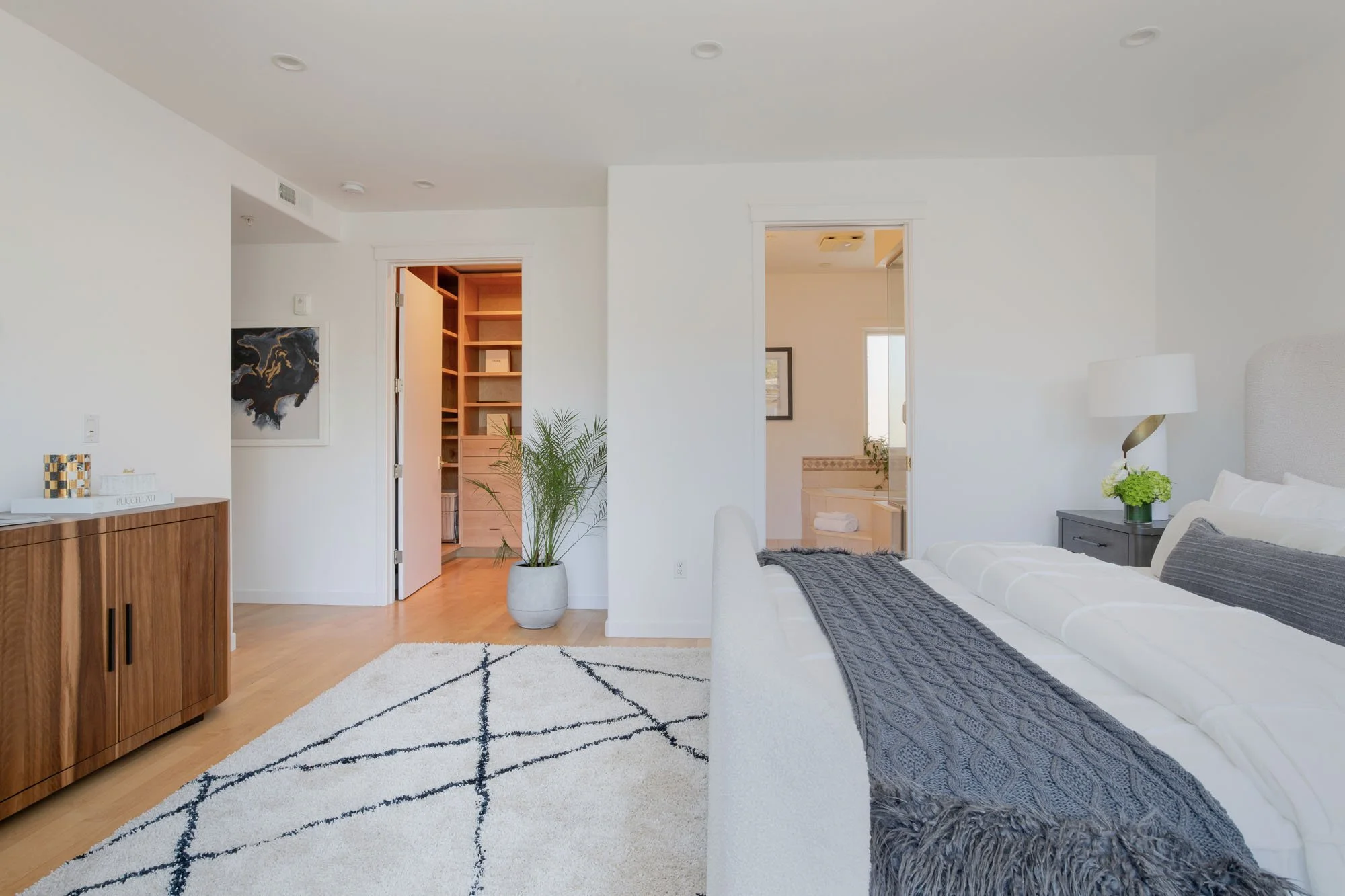 View of a luxury master bedroom featuring a white boucle bed, geometric area rug, and an open doorway leading to a custom walk-in closet with built-in shelving.