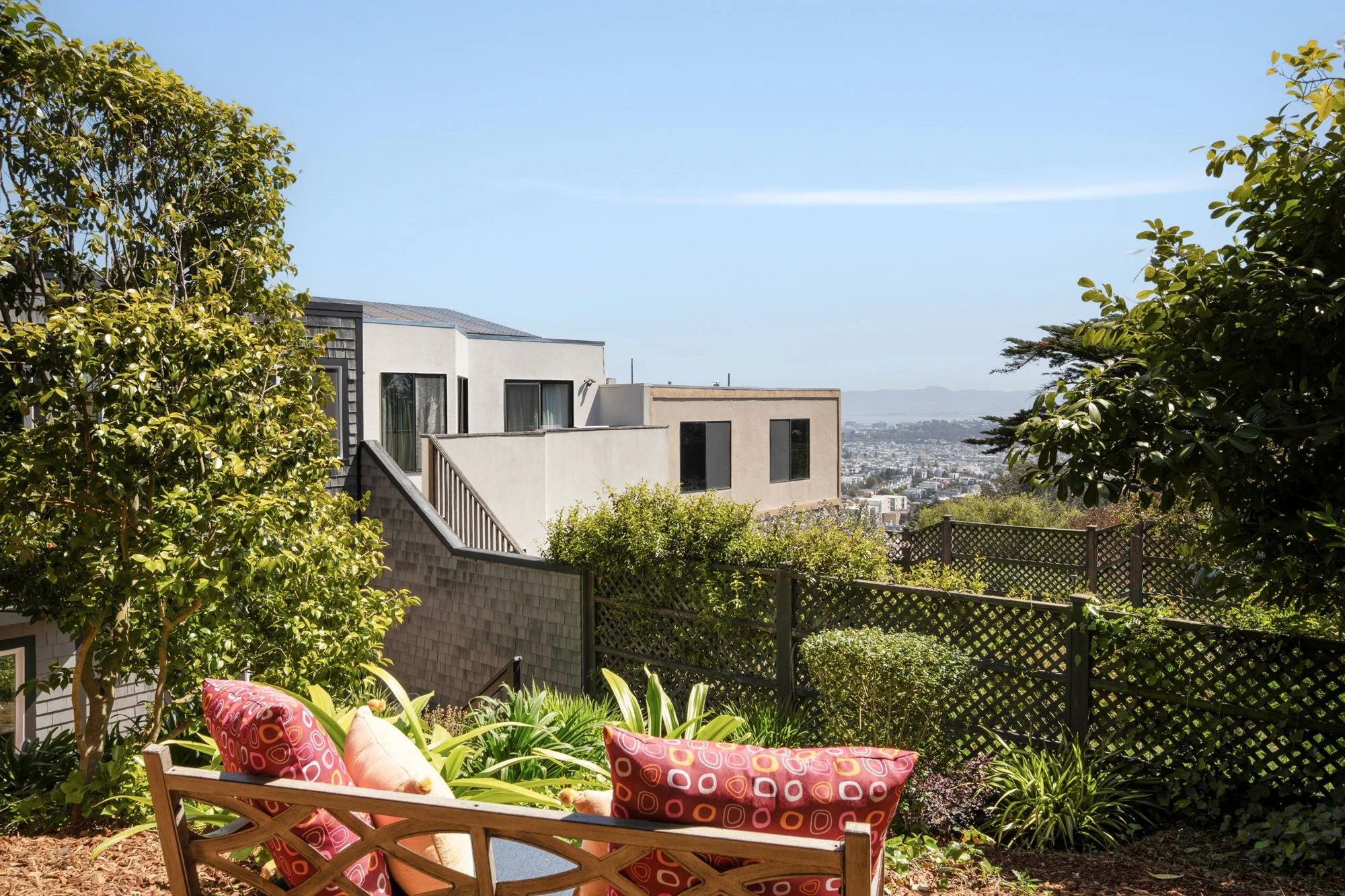 Secluded hillside garden overlook in San Francisco featuring a wooden bench with colorful cushions and a panoramic view of the city skyline and San Francisco Bay.