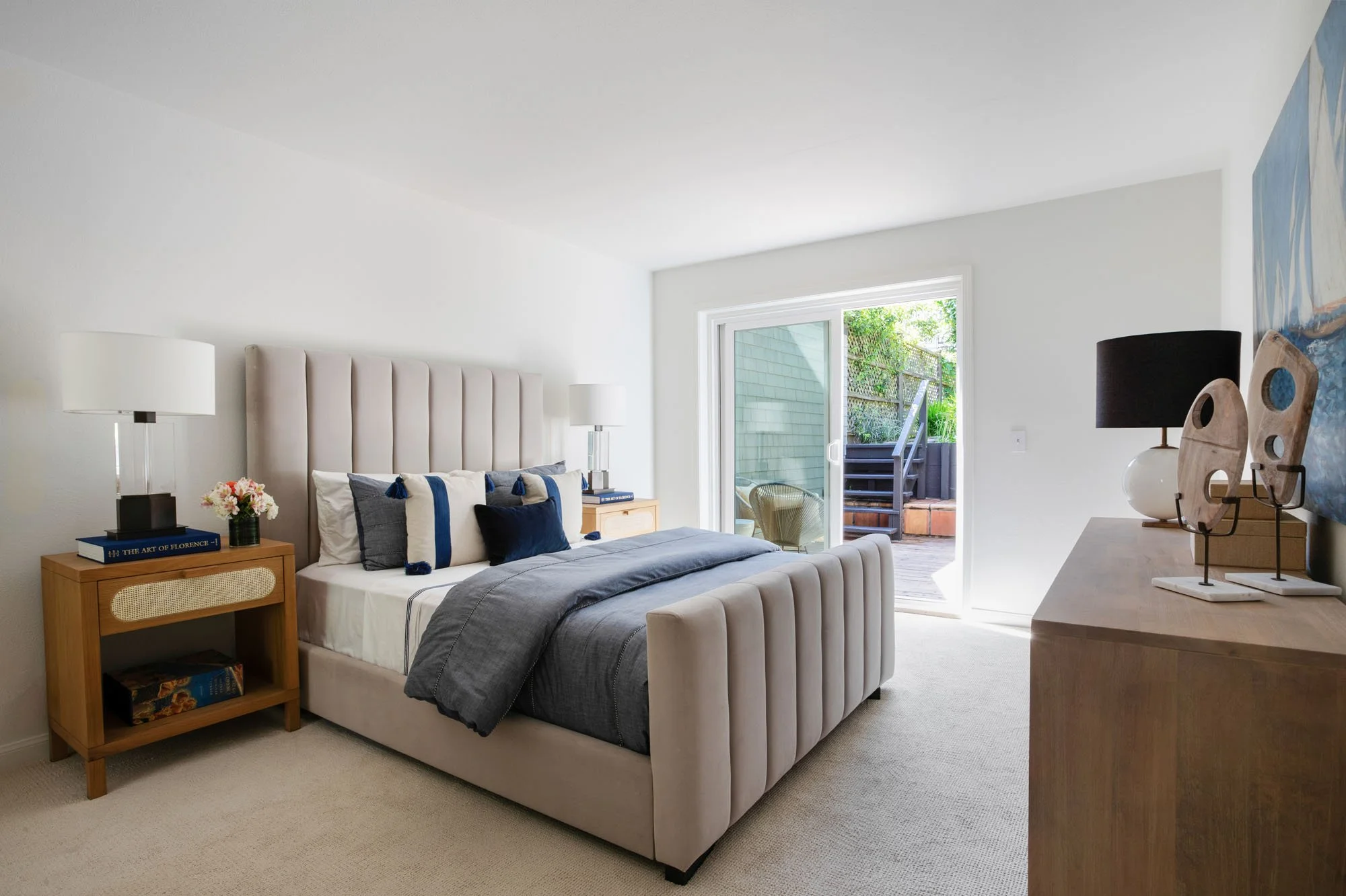 Luxury bedroom suite featuring a beige channel-tufted upholstered bed, coastal blue bedding, and sliding glass doors that lead to a private outdoor deck and terrace.