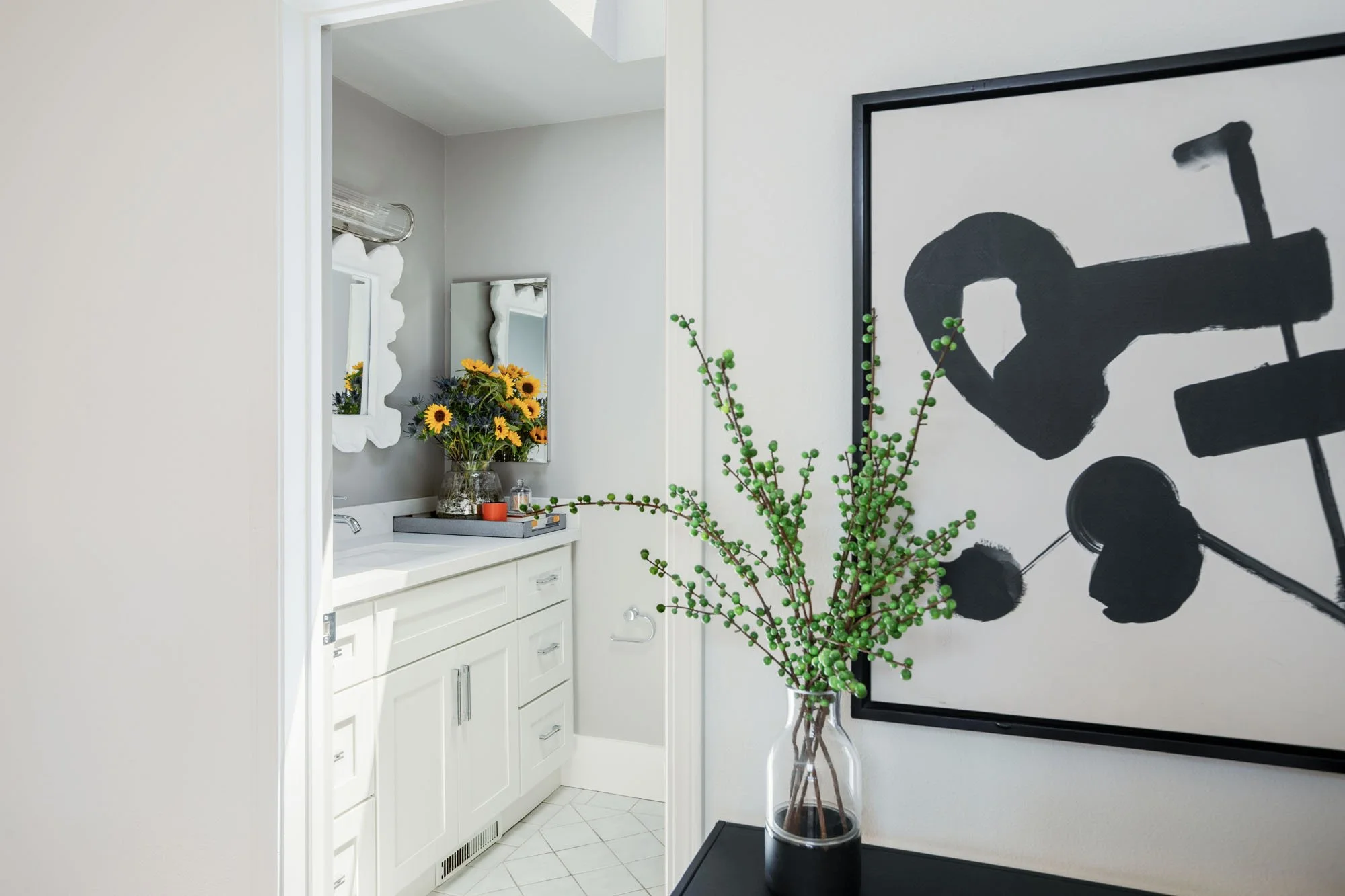 Perspective view of a modern hallway leading to a bathroom, decorated with a large black-and-white abstract painting and a glass vase with green berries.