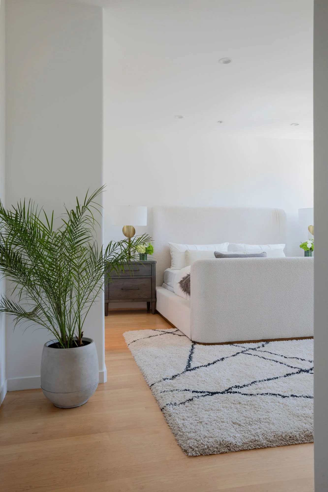 Luxury primary bedroom featuring a white upholstered boucle bed, a geometric shag area rug, and a large indoor palm plant in a grey stone planter.