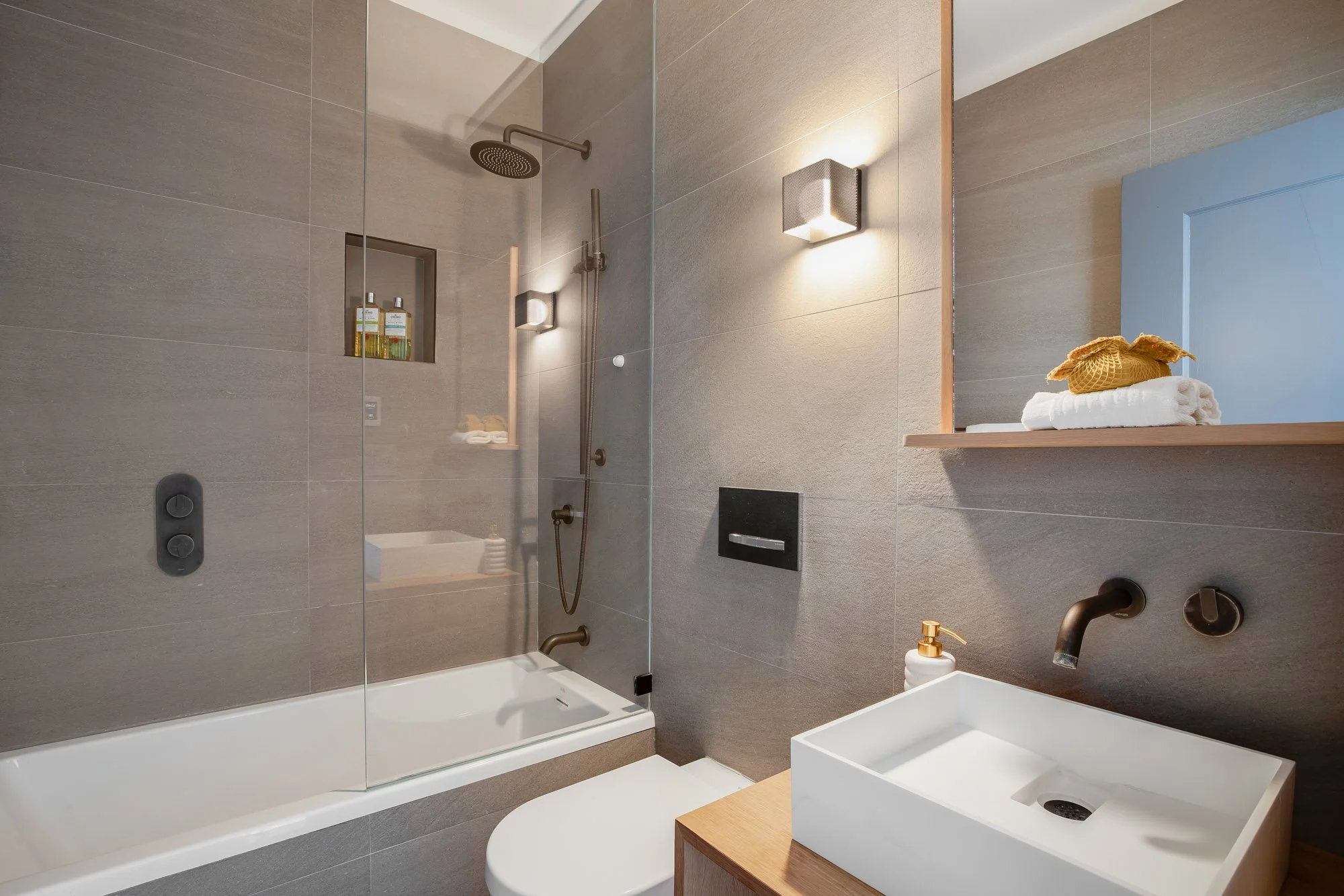 Renovated hallway ensuite bathroom with floor-to-ceiling stone tile, glass-enclosed soaking tub, rain shower head, vessel sink, white oak vanity, and brushed bronze fixtures in luxuy Presidio Heights Victorian condo, San Francisco