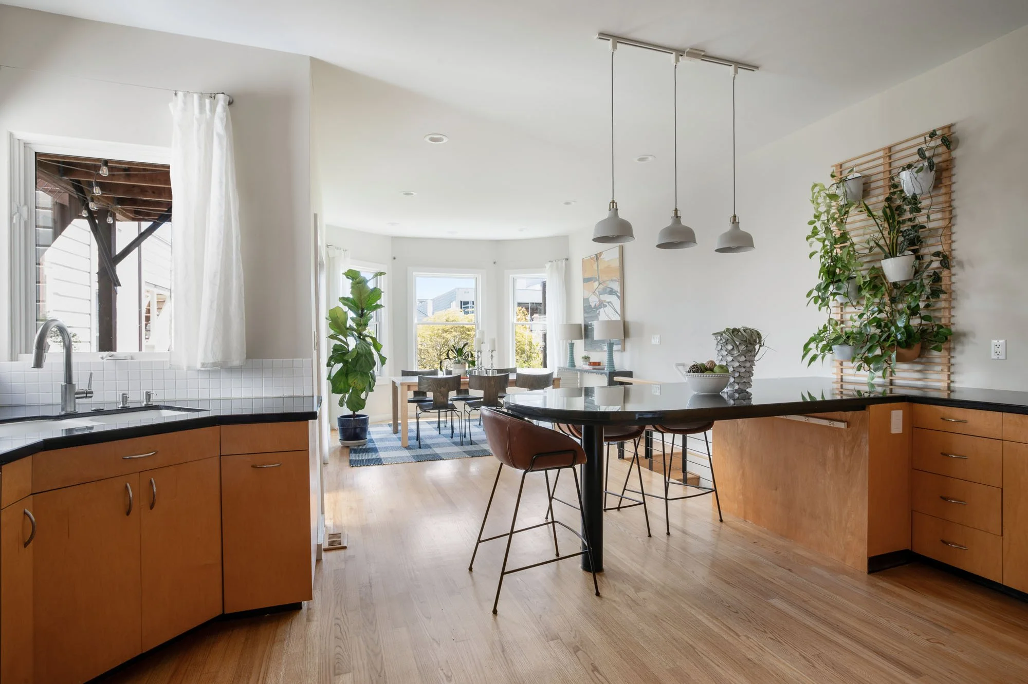 Wide view of an open-plan kitchen and dining area with hardwood floors, white pendant lights, and large windows overlooking the city.
