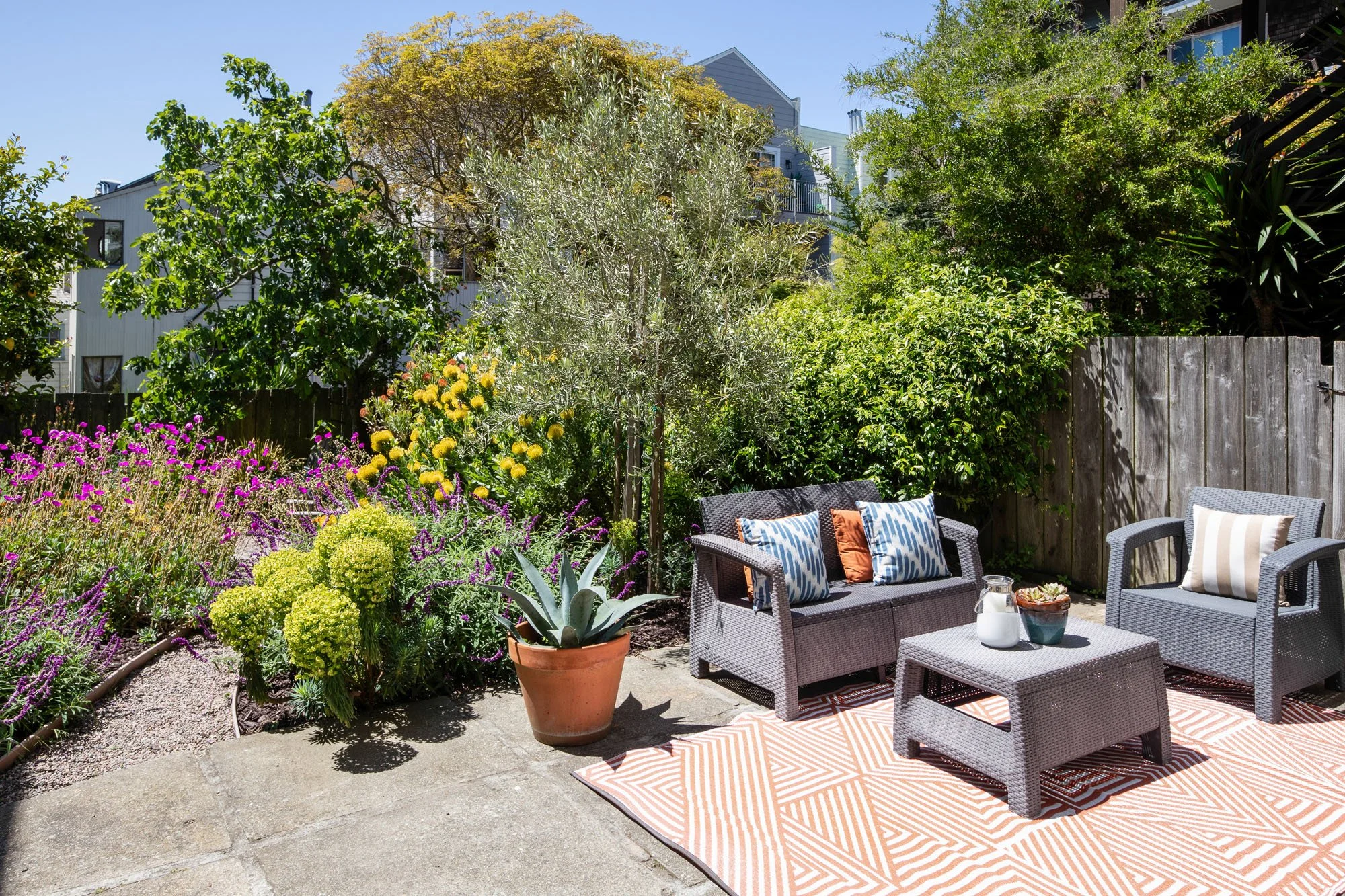 Modern outdoor patio with grey wicker furniture, a geometric orange rug, and a potted agave plant surrounded by a blooming Mediterranean-style garden.