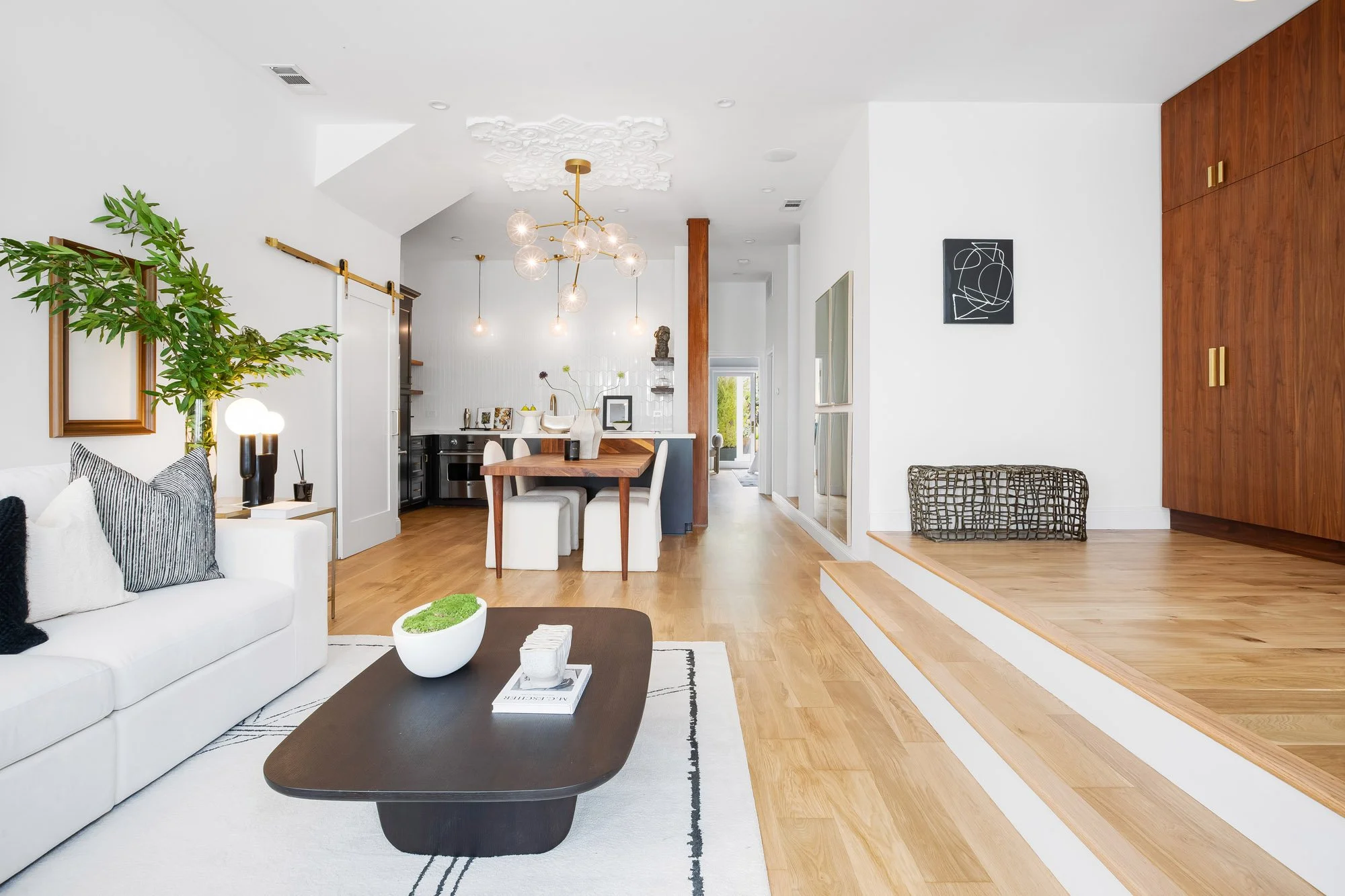 Open-concept living and dining area in San Francisco luxury TIC featuring wide-plank white oak hardwood floors, walnut dining table, gold Sputnik chandelier with ornate ceiling medallion, and custom walnut built-in cabinetry.