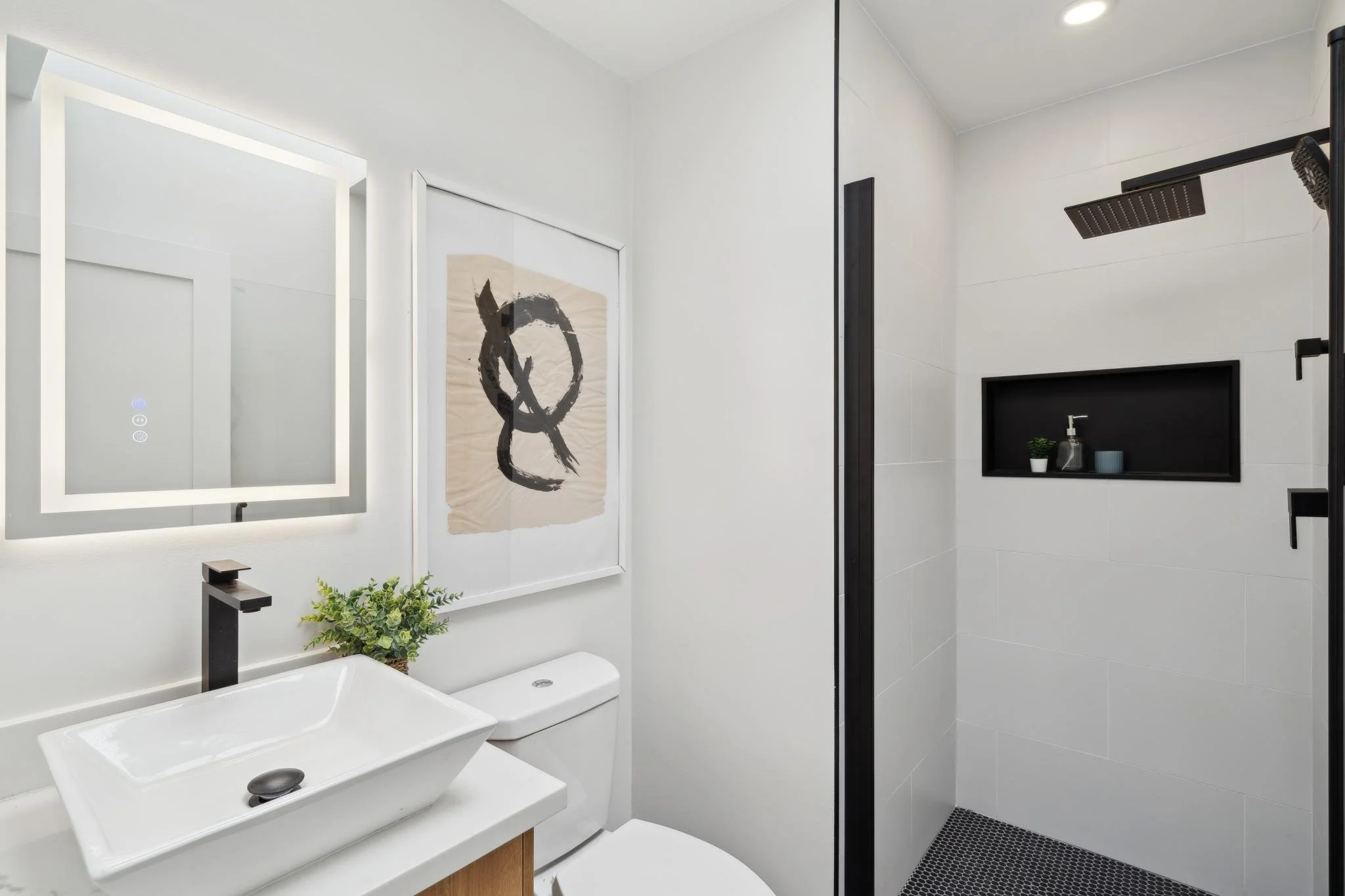 Modern guest bathroom featuring a vessel sink, matte black faucet, back-lit LED mirror, and a walk-in shower with black penny tile flooring.