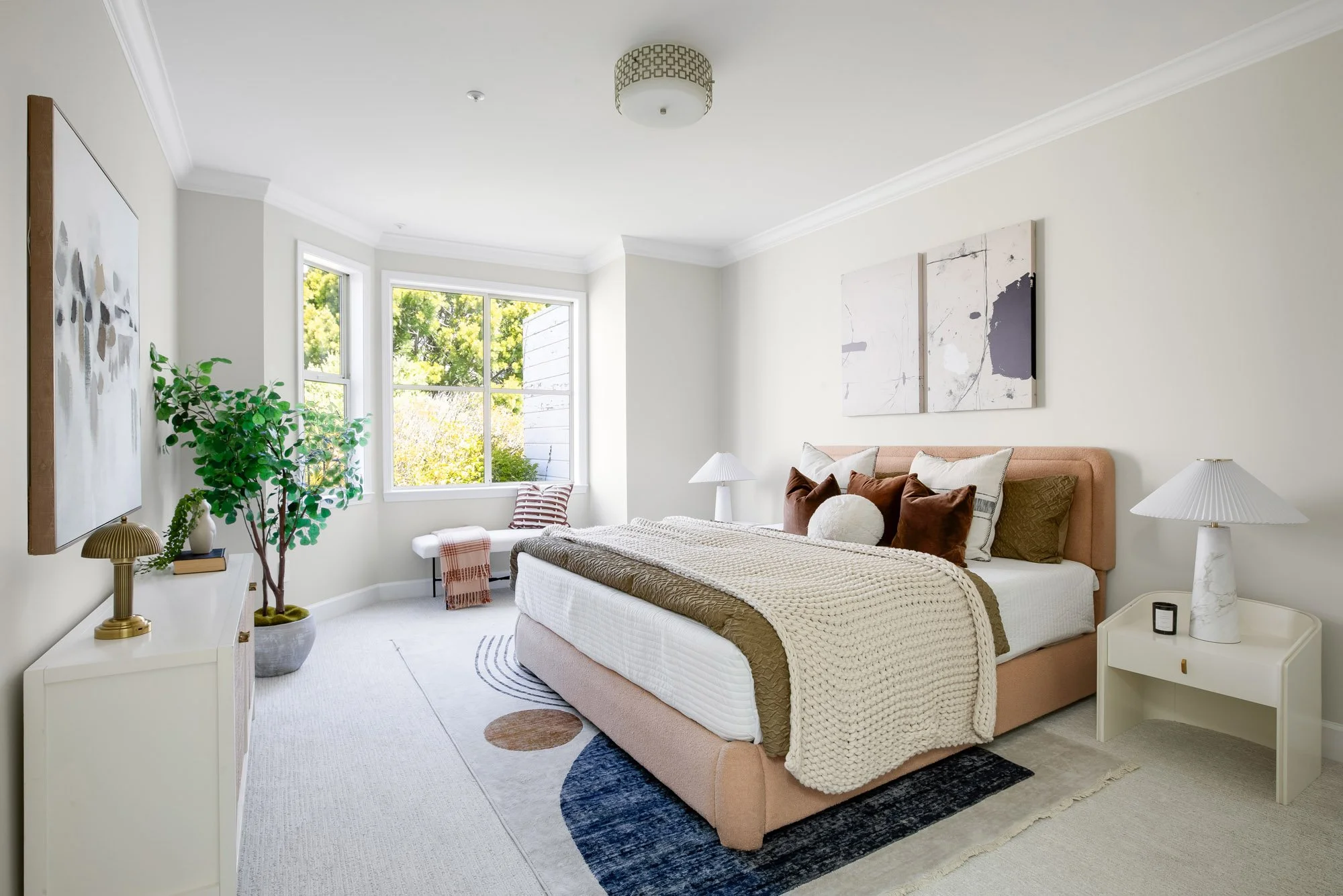 Spacious primary bedroom at 2172 Fell Street San Francisco with bay windows overlooking greenery, blush upholstered king bed, carpet flooring, abstract wall art, and white dresser with brass lamp