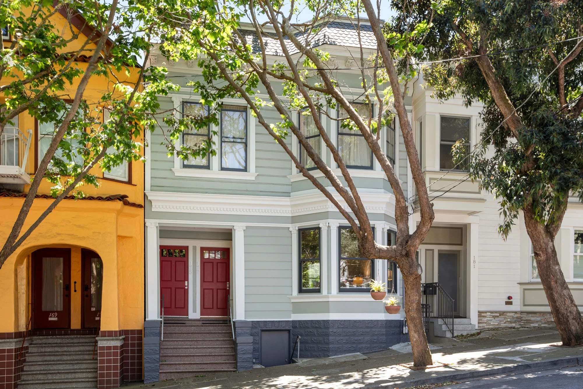 Classic San Francisco Edwardian-style duplex with a light green wood-sided exterior, red double front doors, and large bay windows on a tree-lined street.