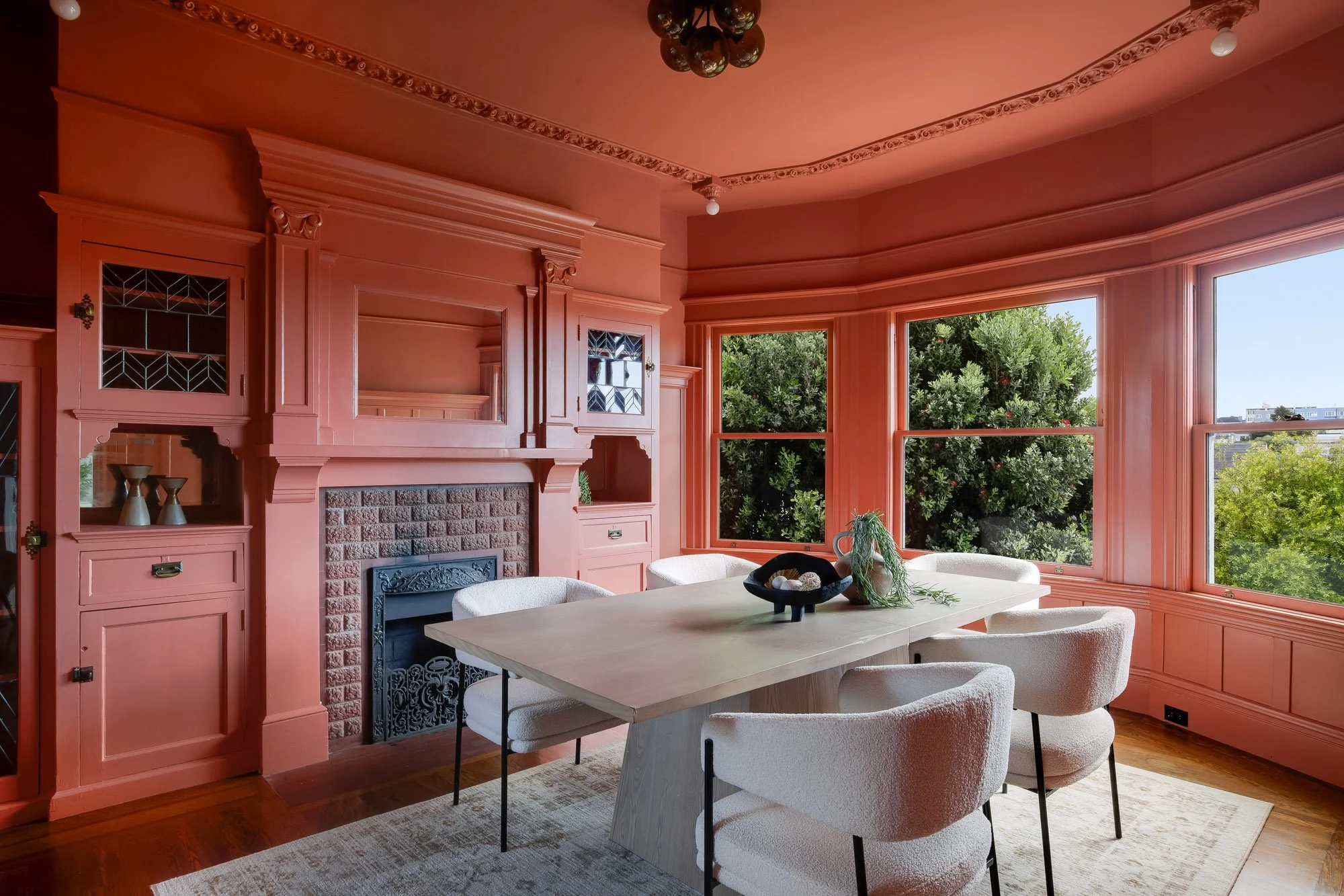 Dramatic Victorian formal dining room with floor-to-ceiling terracotta paint, original ornate ceiling molding, leaded-glass built-ins, decorative fireplace, curved bay windows with treetop views, Presidio Heights, San Francisco