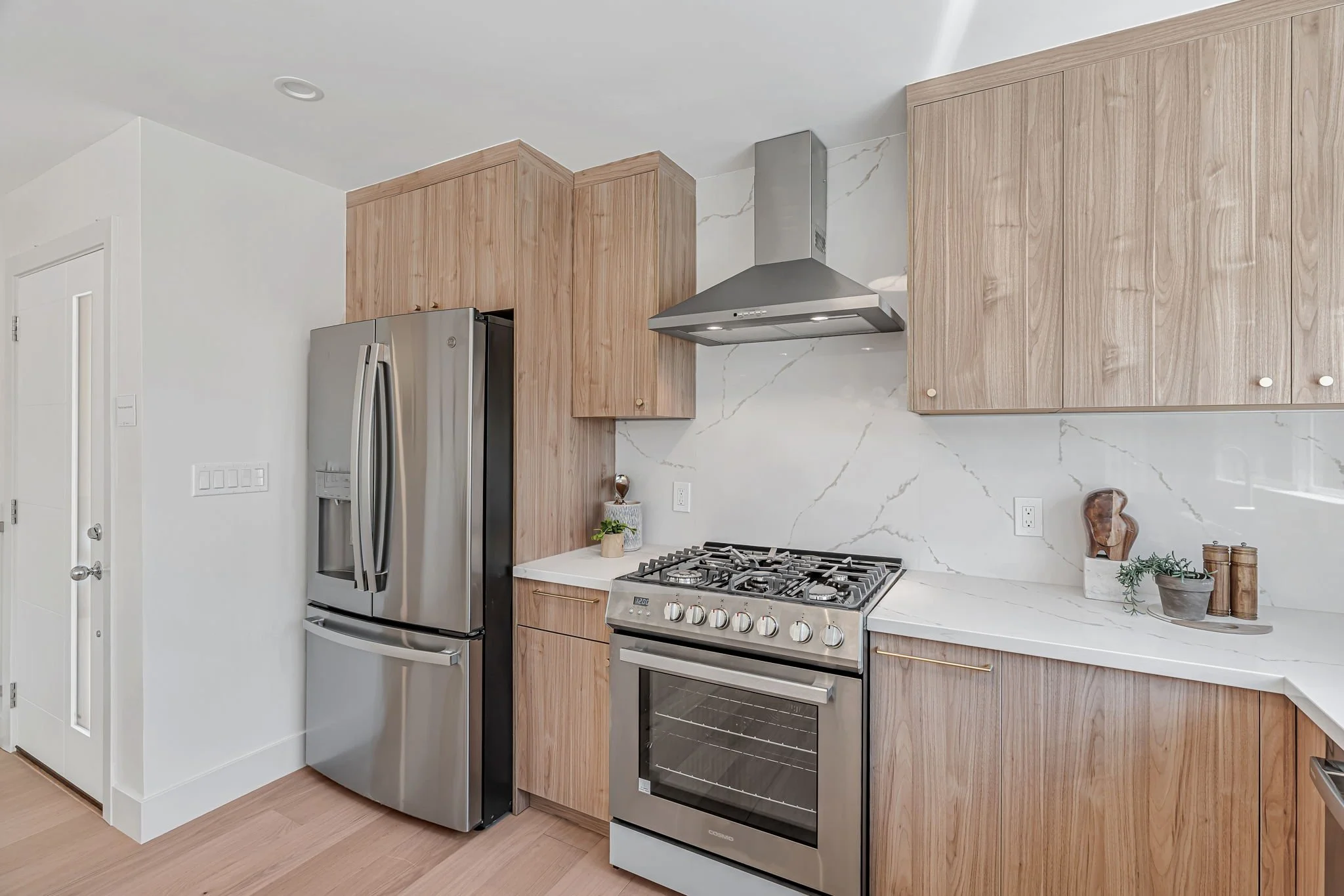 A focused shot of the kitchen’s stainless steel workspace. It features a professional-grade gas range with a chimney hood and a large French-door refrigerator, all framed by seamless white marble-look quartz countertops and light wood cabinets.