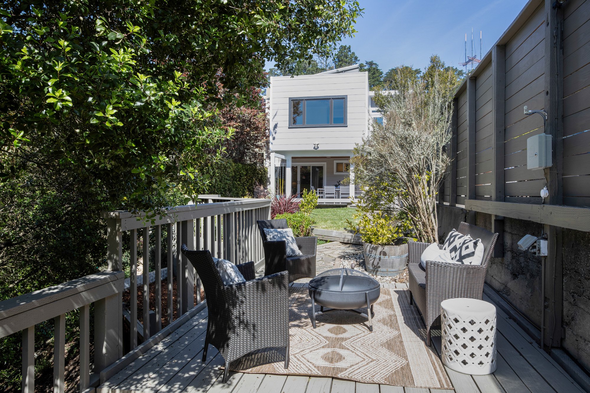 Modern outdoor living space with wicker chairs, fire pit, and patterned rug on a gray wooden deck overlooking a lush backyard.