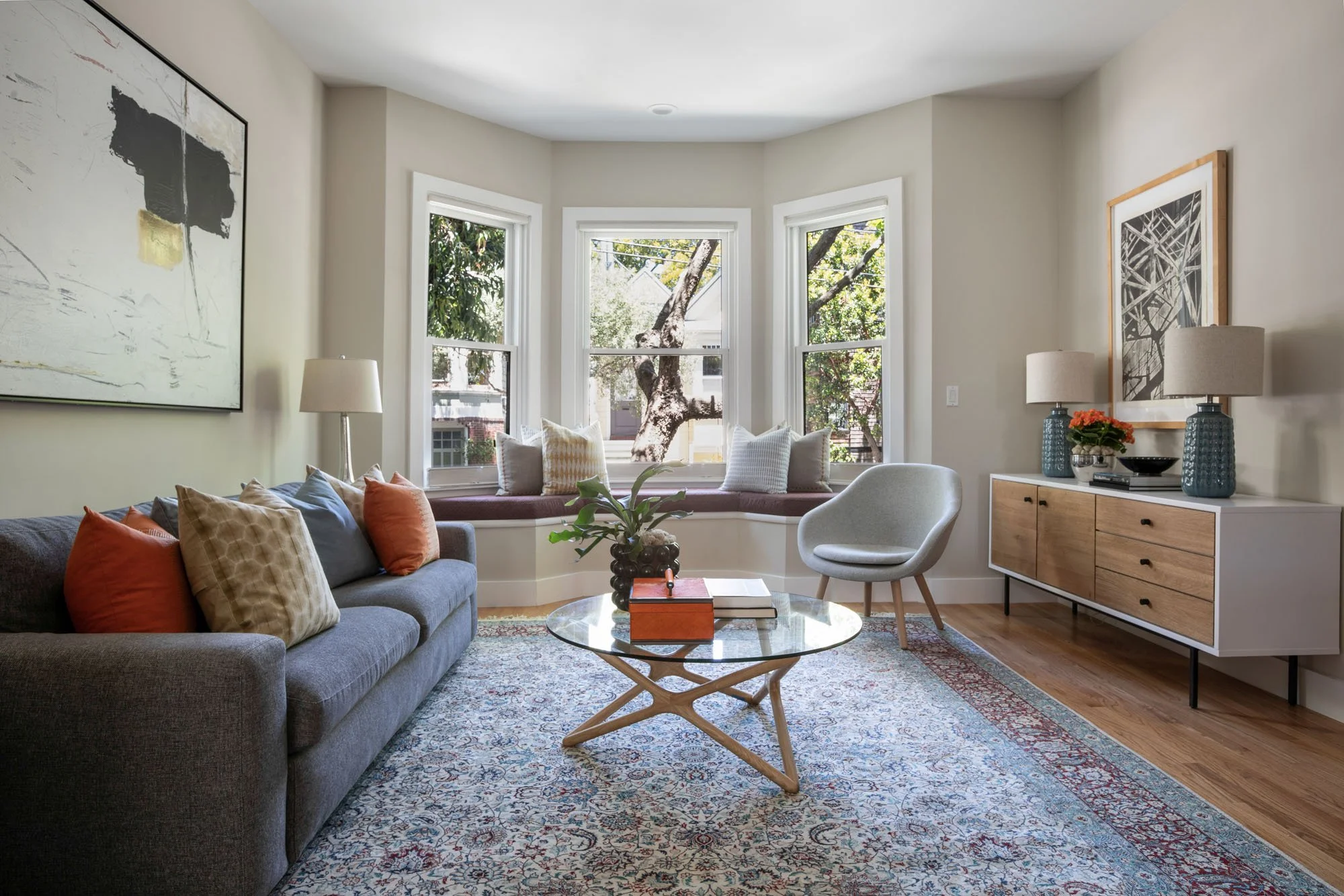 Sunlit living room featuring a curved bay window with a purple cushion seat, a mid-century modern sideboard, and a grey sofa with orange accent pillows.