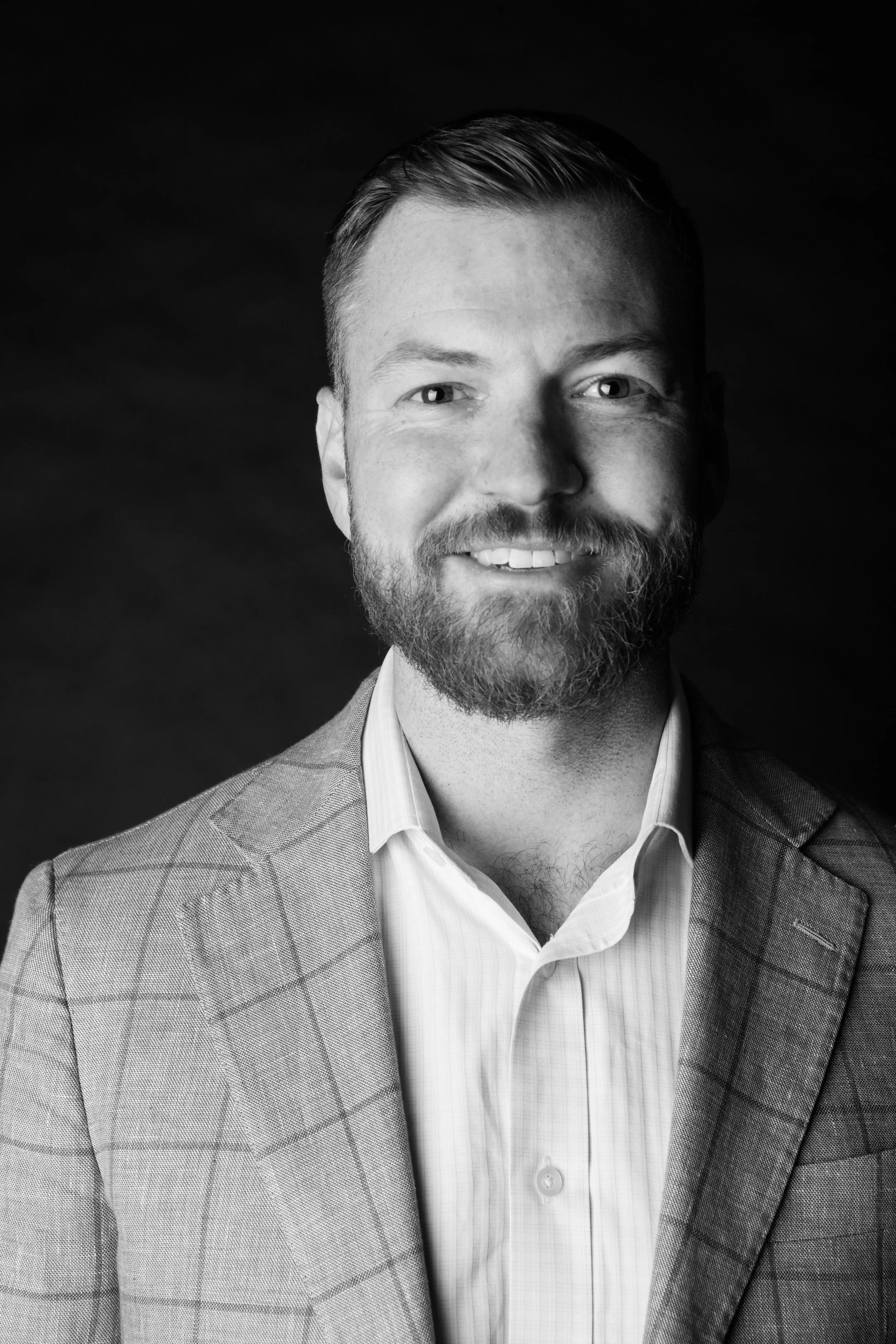 A black and white portrait of a smiling man with a beard, wearing a checkered blazer and a light-colored shirt.