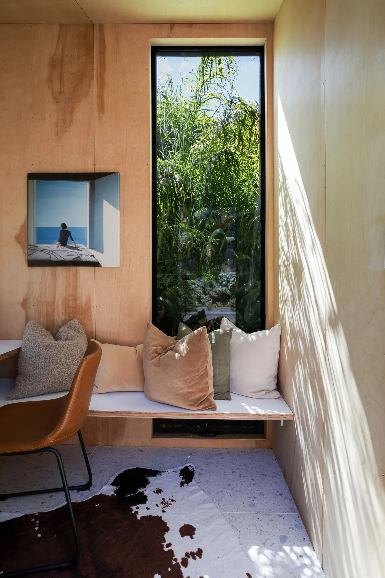 Interior detail of the garden office showing a built-in plywood bench with plush neutral pillows and a large vertical window overlooking the lush backyard.
