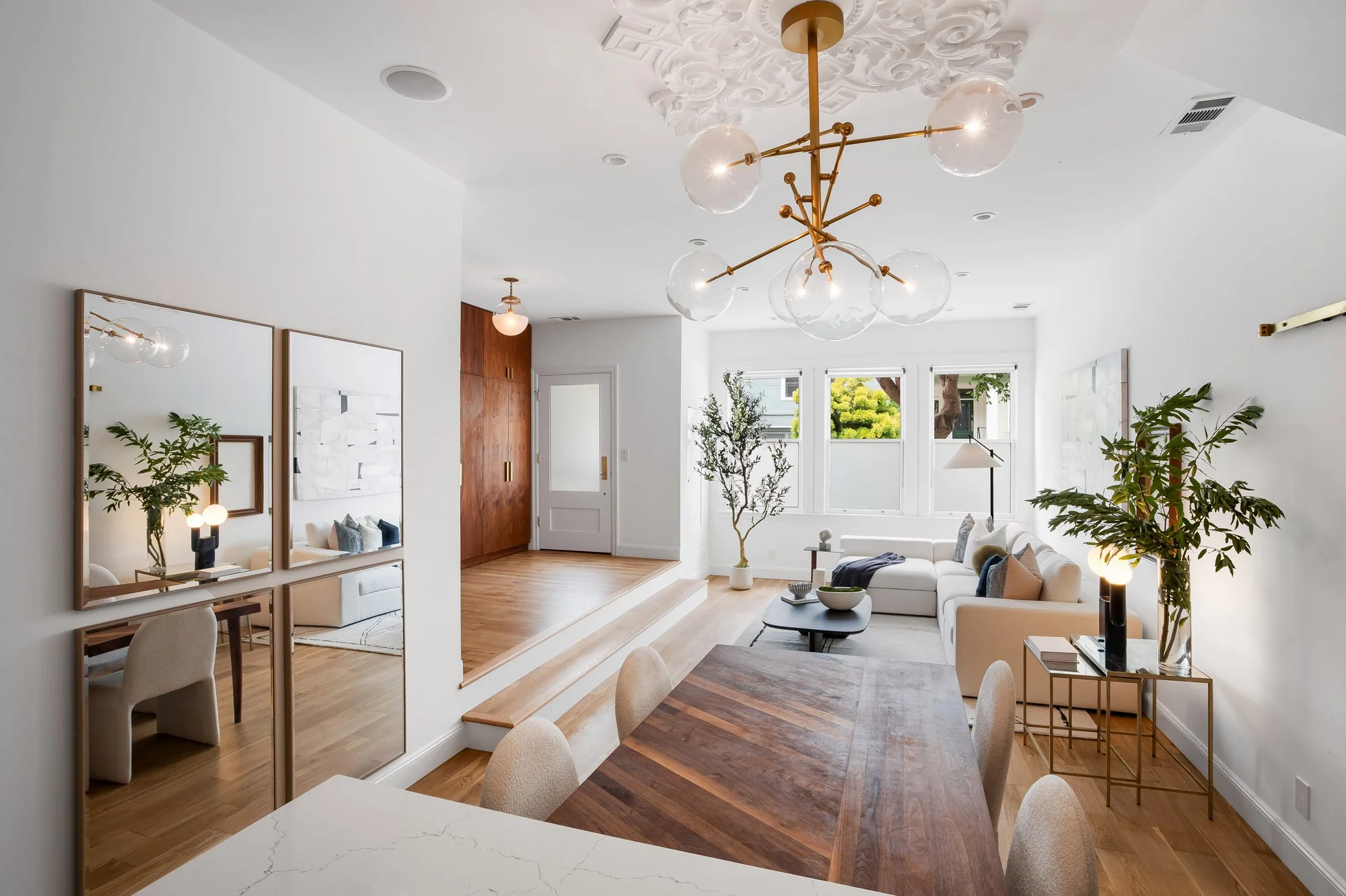 Expansive open-plan great room in Castro San Francisco luxury TIC showing quartzite kitchen island, walnut dining table, stepped platform living area, ornate plaster ceiling medallion, brass Sputnik chandelier, and street-facing windows