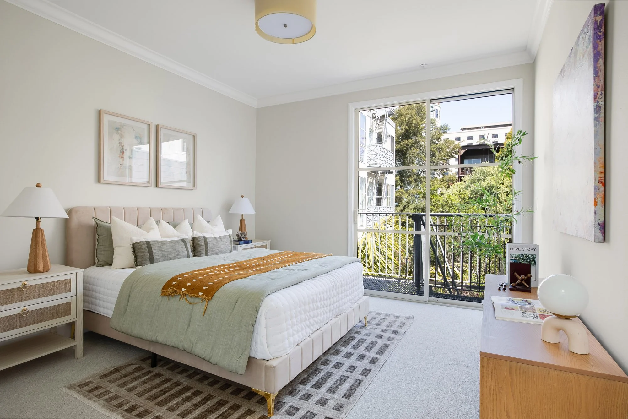 Sunlit second bedroom at 2172 Fell Street San Francisco with private balcony access, large sliding glass door with tree and city views, tufted upholstered queen bed, rattan nightstands, carpet flooring, and crown molding