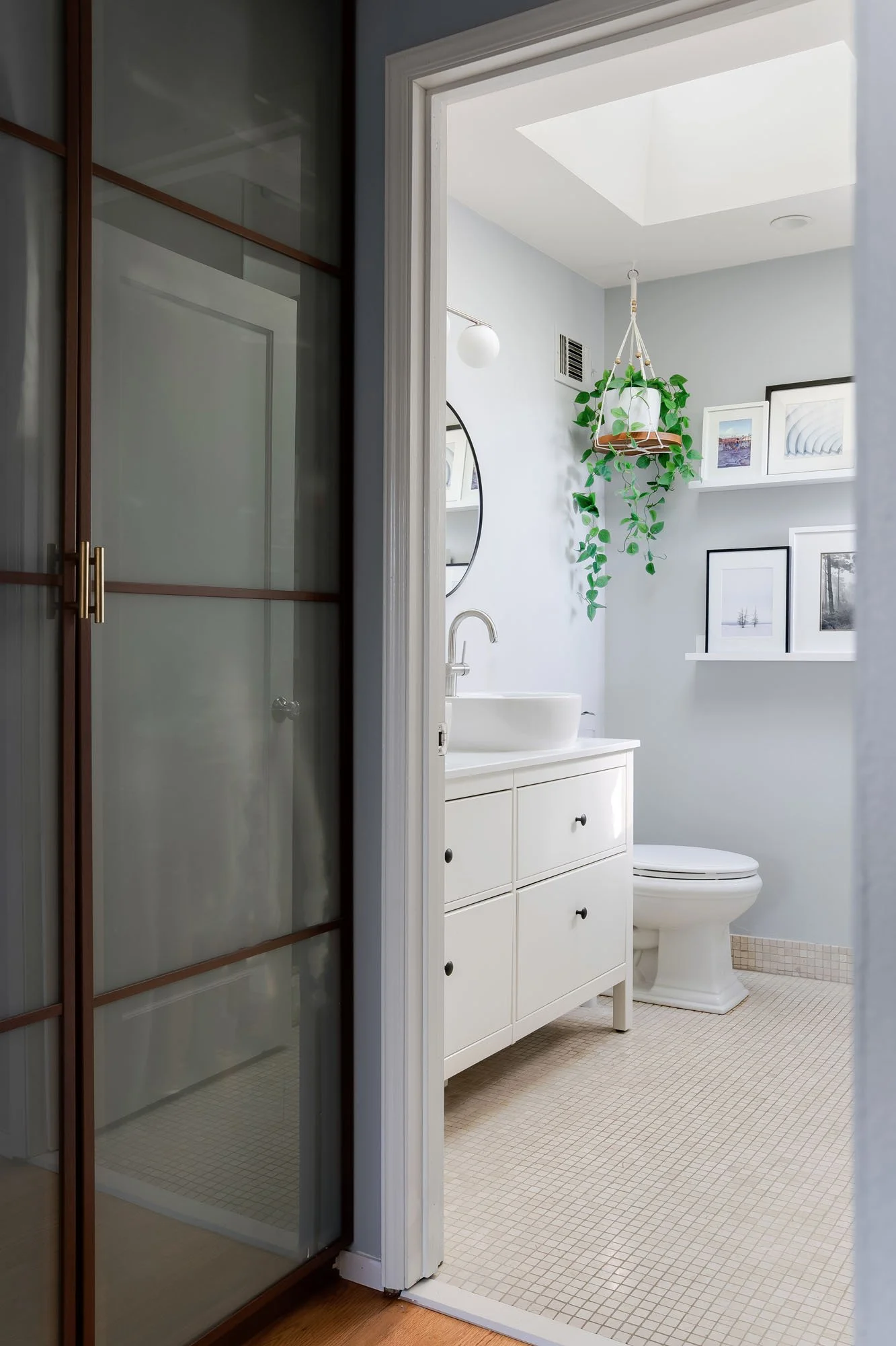 Bright ensuite bathroom featuring a white vanity with vessel sink, round black mirror, and a hanging plant under a skylight.