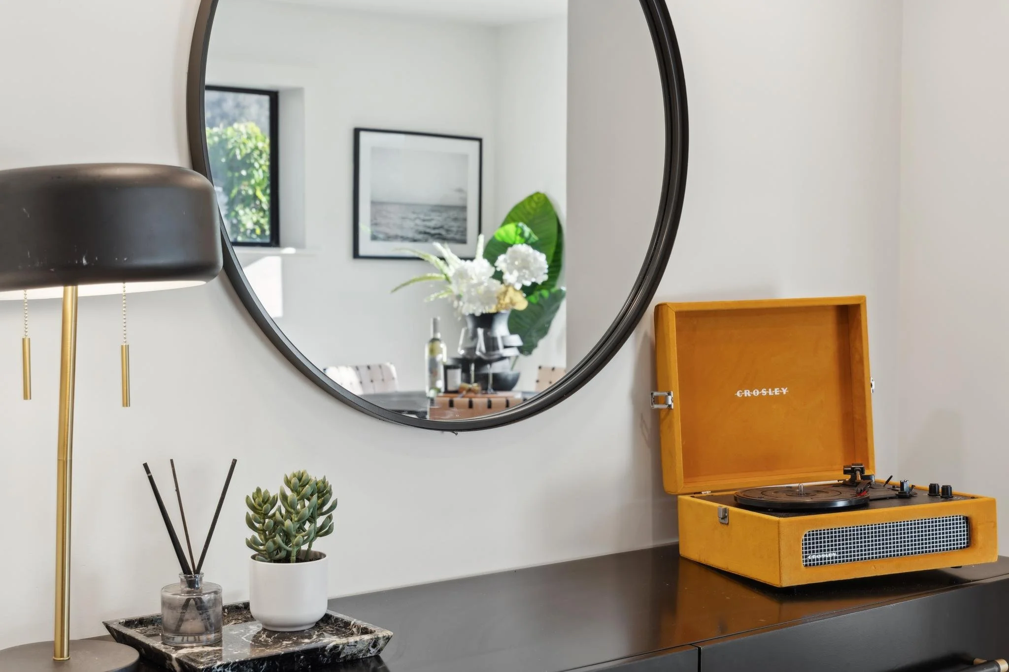 Close-up of interior styling featuring a mustard yellow record player, matte black lamp, and a large circular mirror reflecting a sunlit dining area.