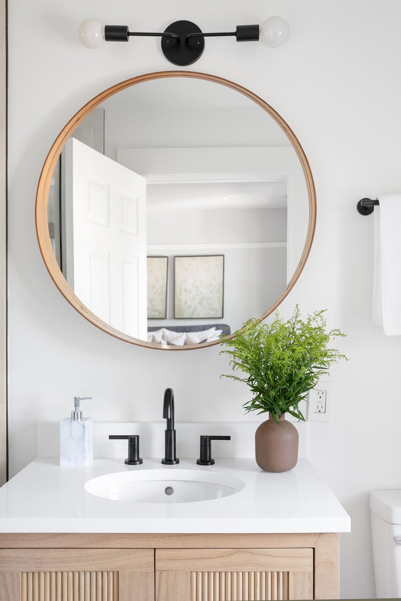Close-up of a modern bathroom vanity featuring a light wood cabinet with slatted details, a white countertop, matte black fixtures, and a large round wood-framed mirror reflecting the primary bedroom.