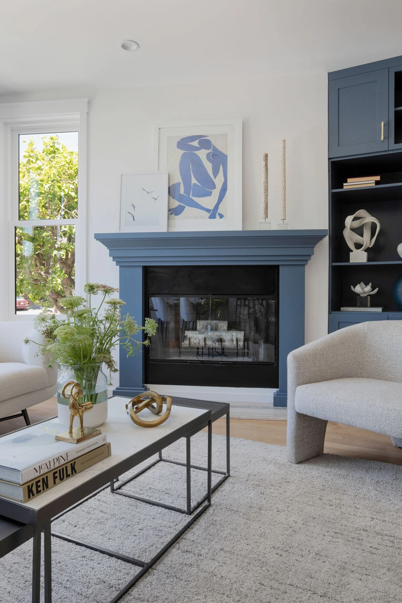 Close-up of a blue fireplace mantle with minimalist decor, a white textured accent chair, and a multi-tiered black coffee table on a grey shag rug.