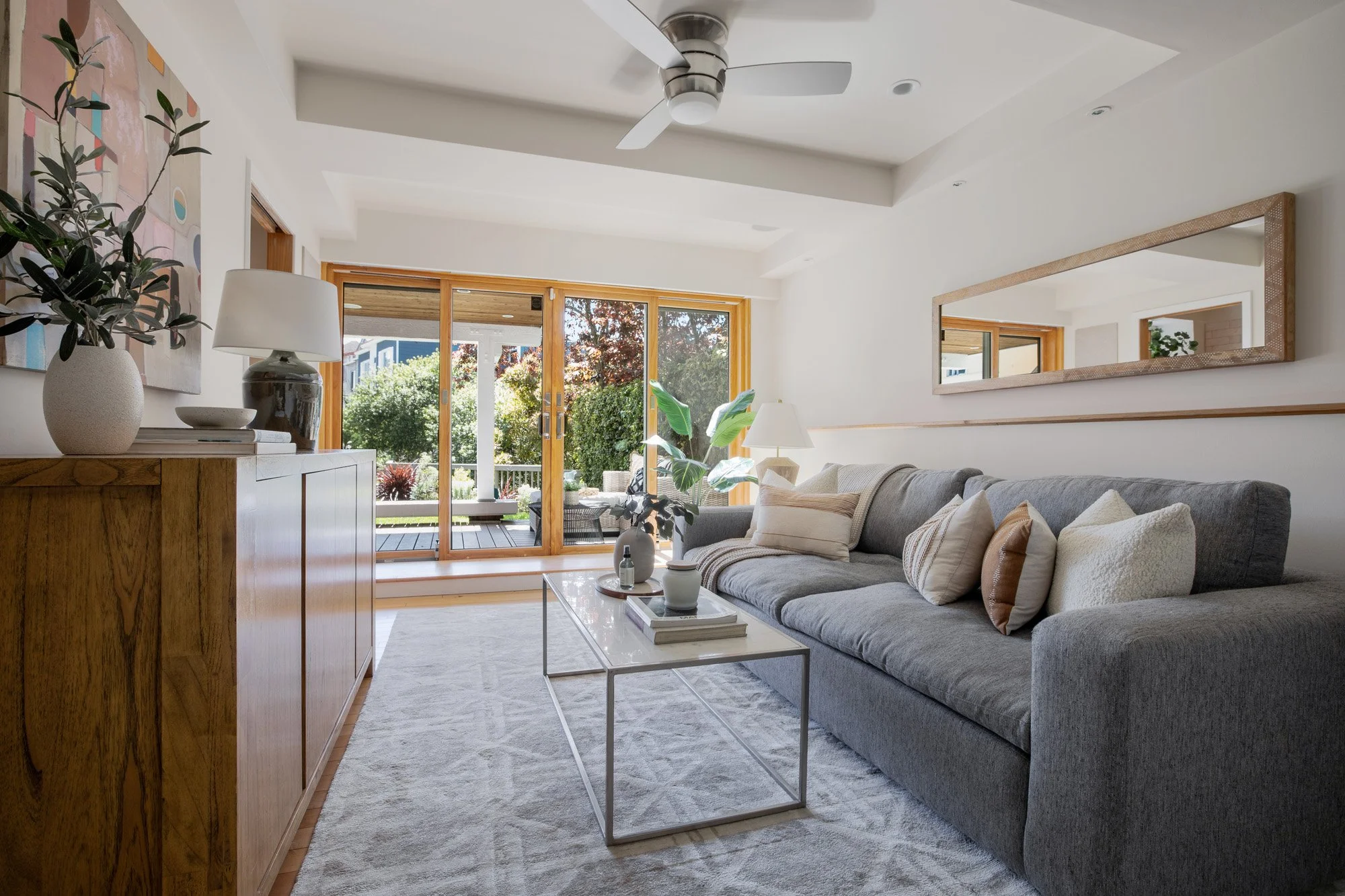 Sun-drenched modern living room featuring a large gray sectional, minimalist coffee table, and floor-to-ceiling glass doors leading to the garden.
