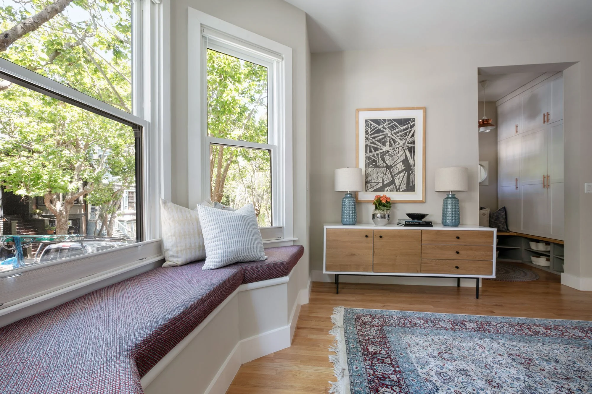 Detail view of a built-in window bench next to a wood and white console table with blue lamps, featuring hardwood floors and neutral walls.