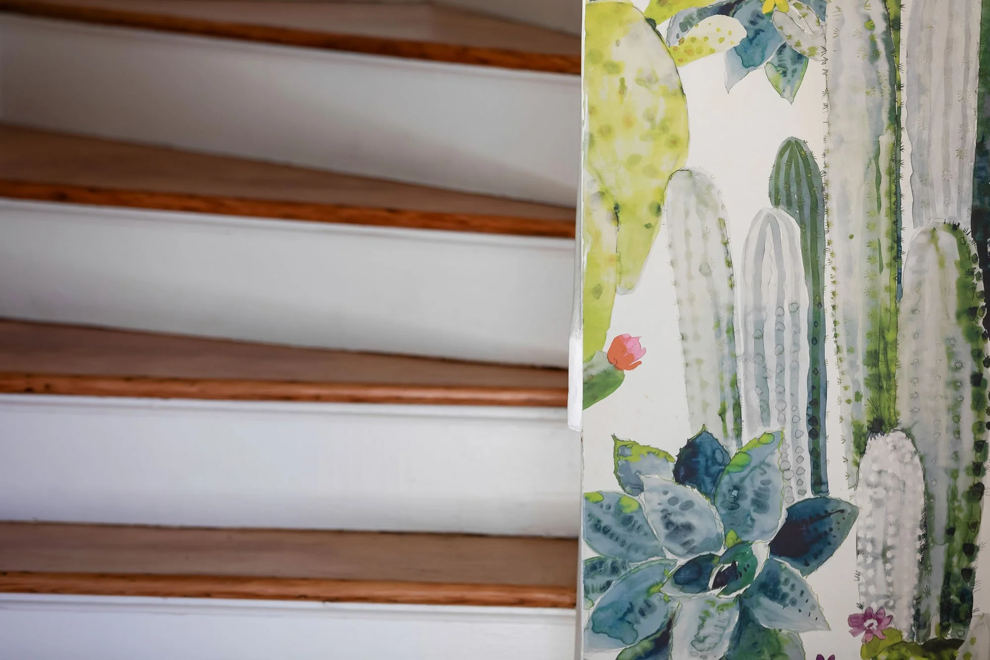 Artistic detail of a white staircase next to a vibrant wallpaper featuring a watercolor cactus and succulent motif.