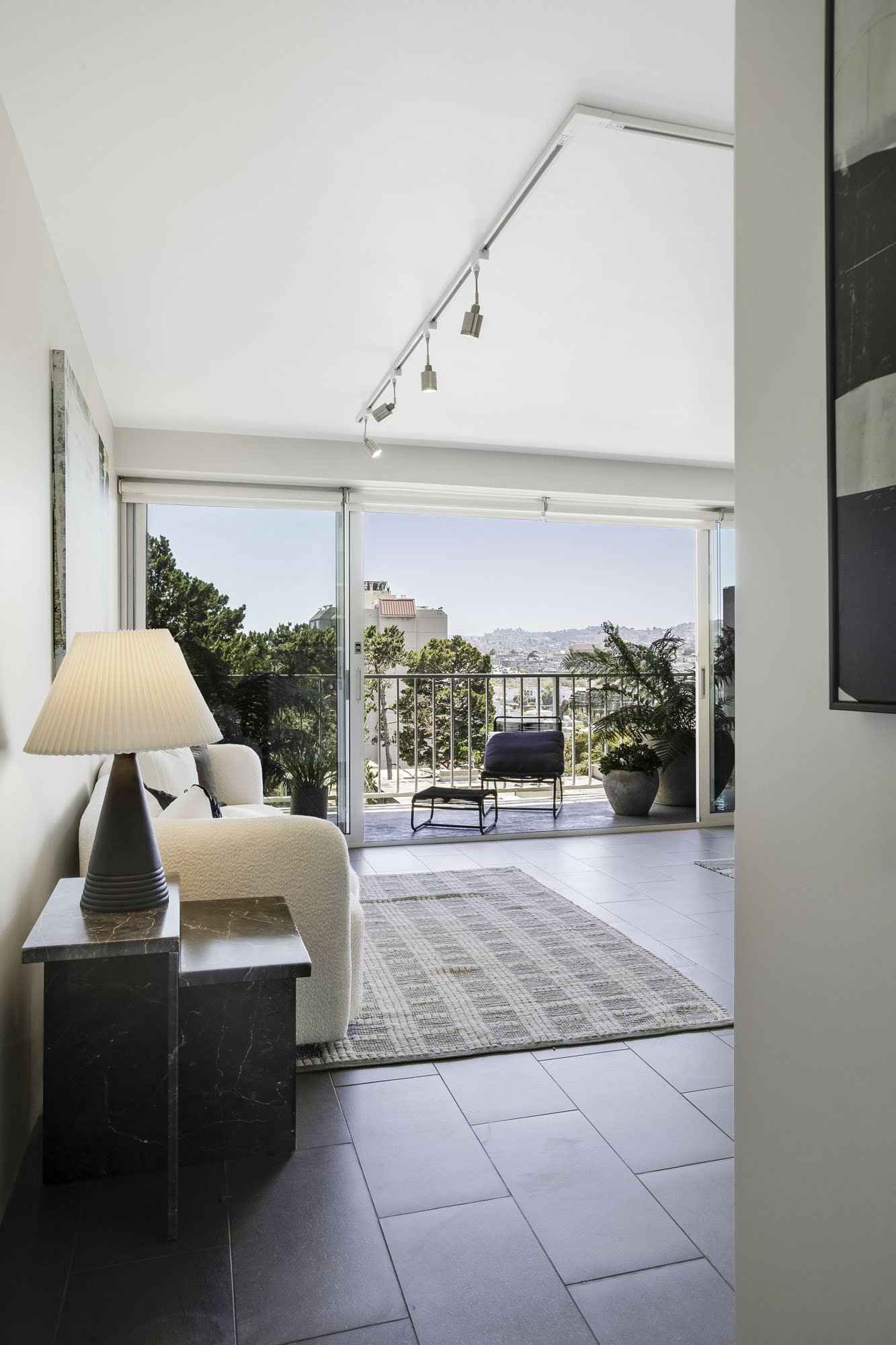 Modern apartment living room in 66 Cleary #601, featuring dark tile floors, a white sofa, and floor-to-ceiling glass doors opening onto a balcony with city and mountain views.