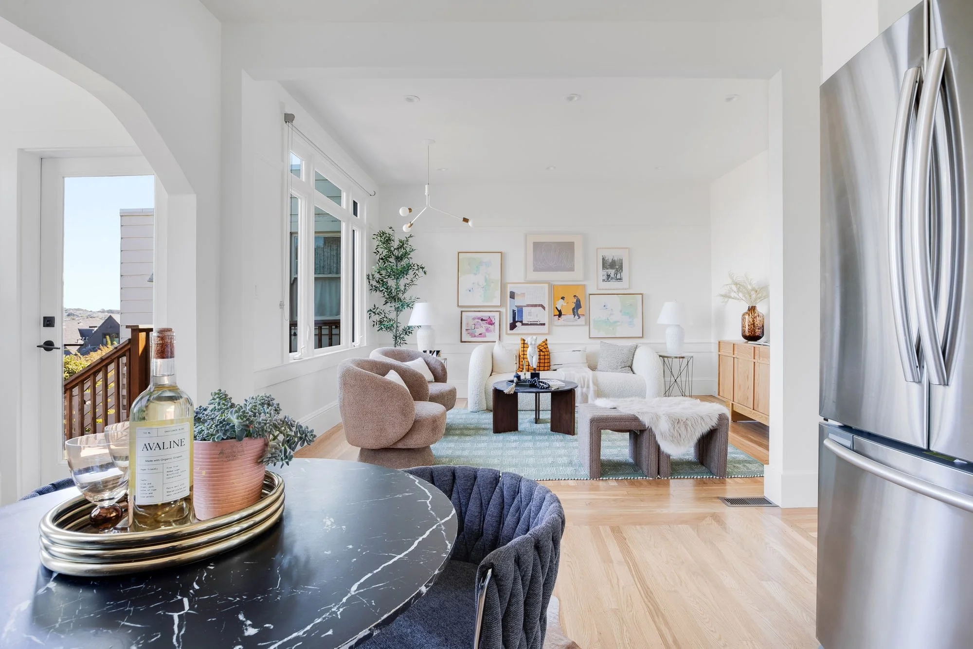View from the dining area into the spacious living room, showcasing a gallery wall of colorful art, plush boucle armchairs, and a large window overlooking the deck.