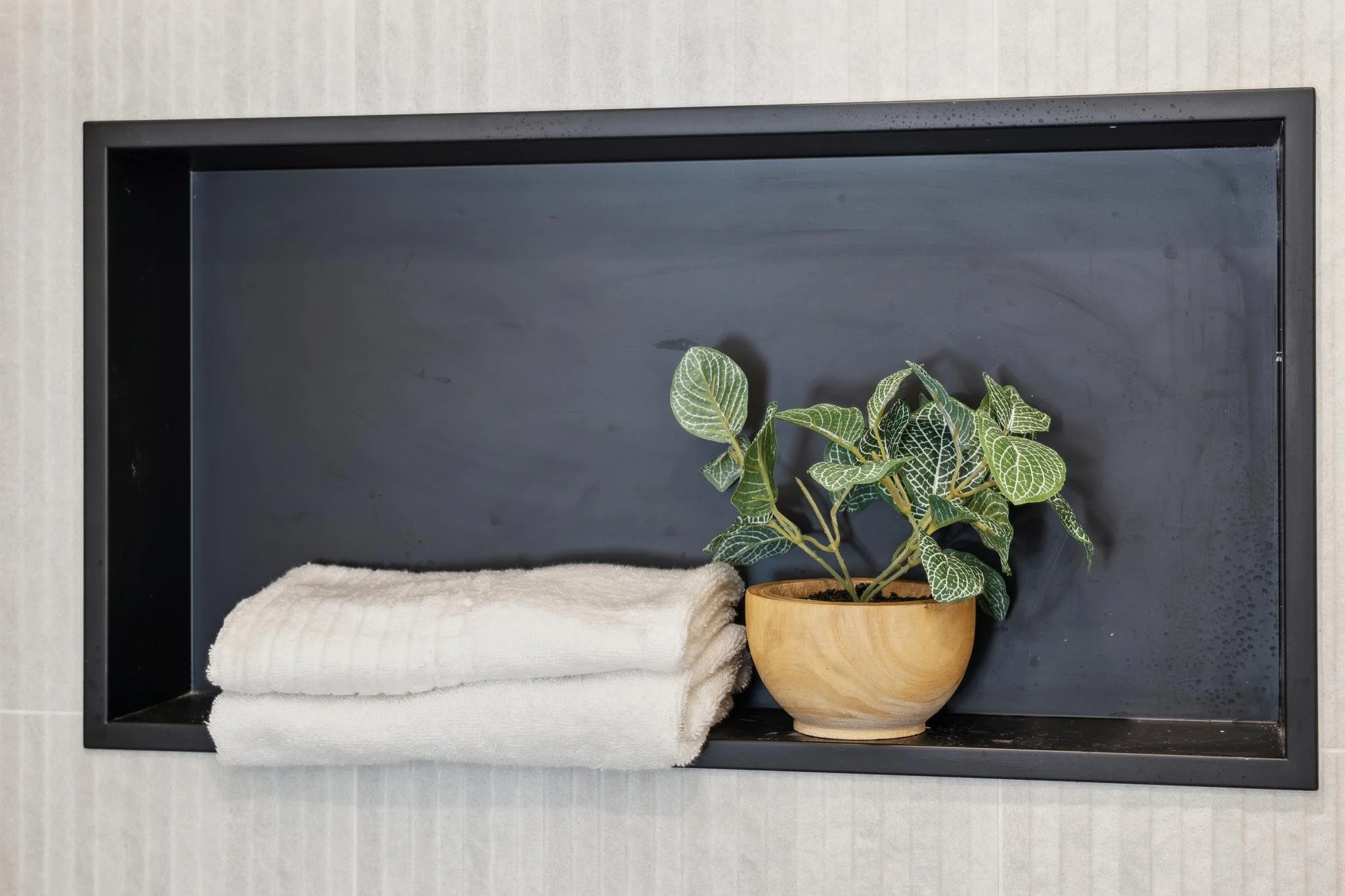 Detail shot of a black recessed shower niche featuring a small potted plant and folded white towels against textured white tile.
