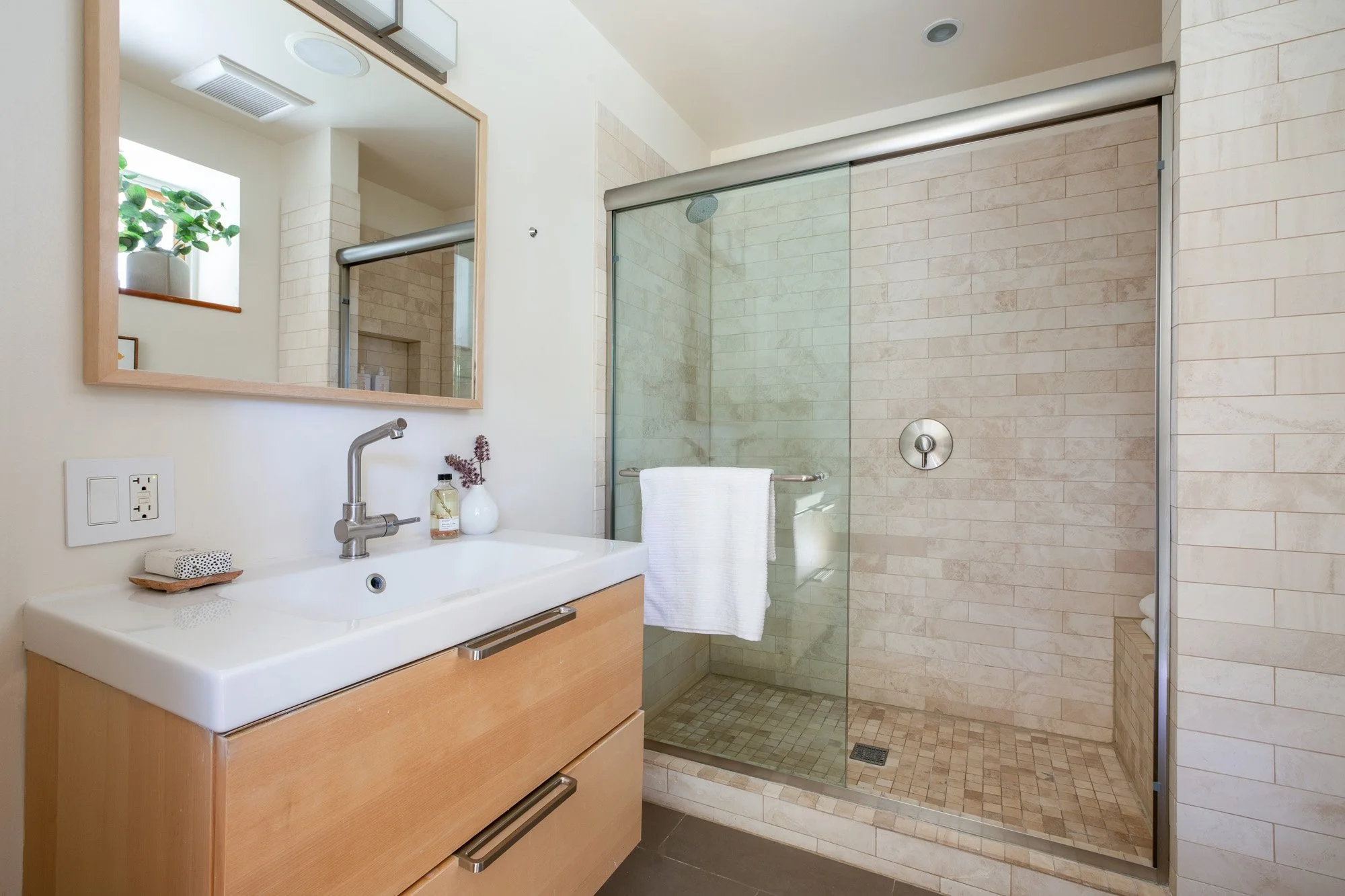 Renovated bathroom with a light wood vanity, white vessel sink, and a walk-in shower with tan subway tile.