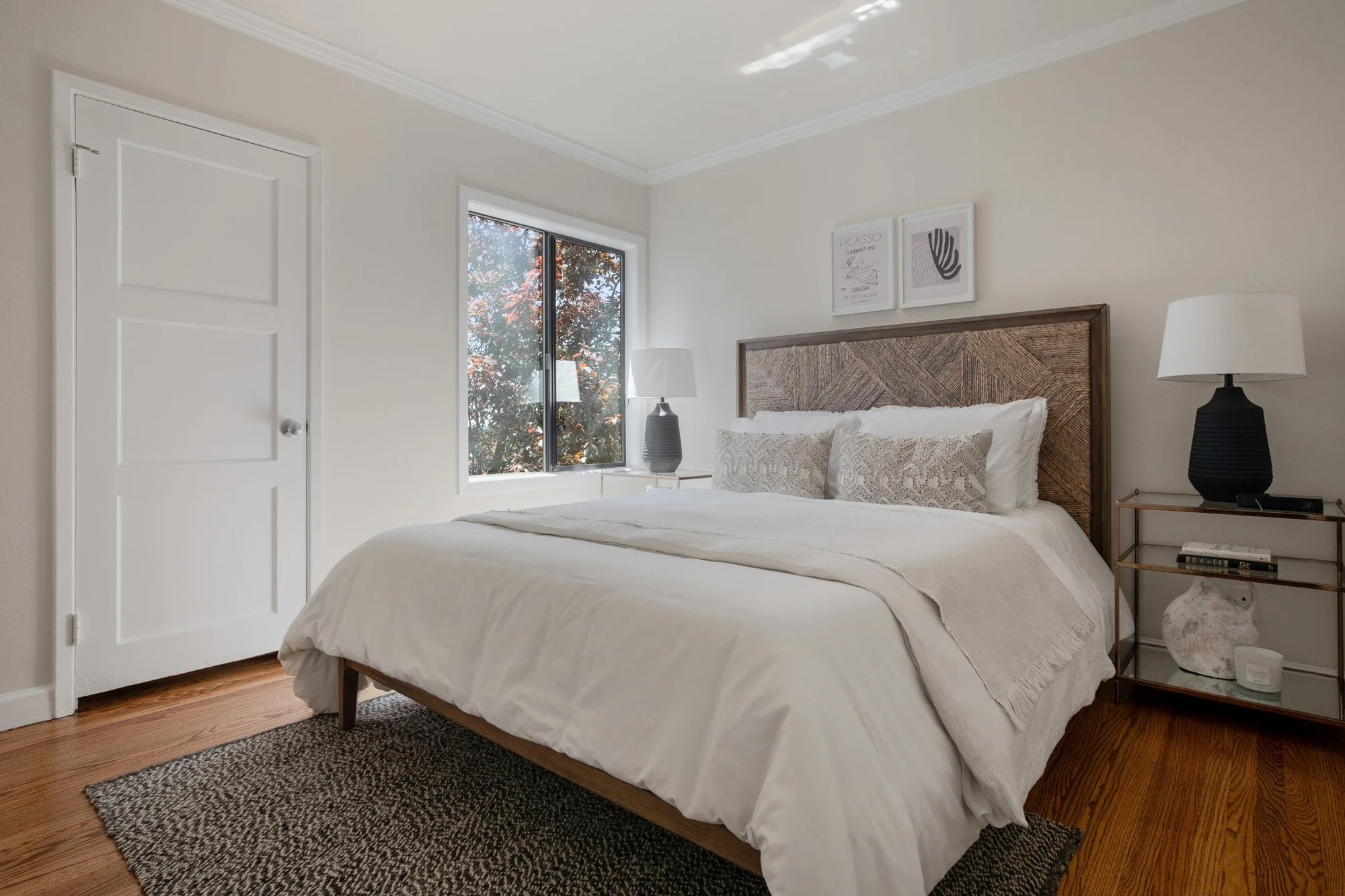 Primary bedroom with a woven wood headboard, white linens, hardwood floors, and a large window providing natural light.