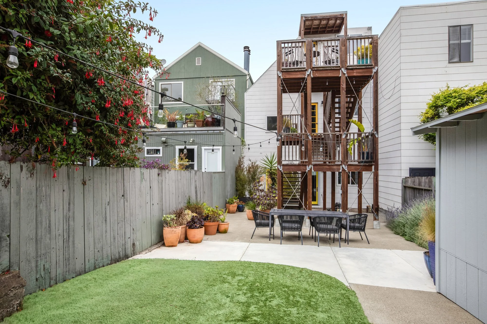 Full view of shared rear garden at 4545 18th Street Castro San Francisco TIC showing artificial turf lawn, concrete patio, black rope outdoor dining set, multi-level redwood deck and staircase, string lights, fuchsia-covered fence, and yellow unit en