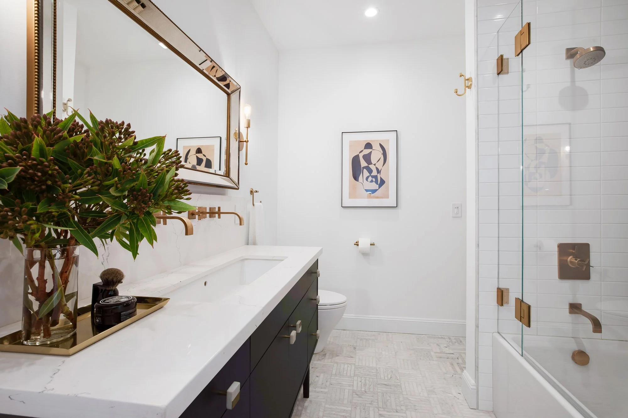 Renovated secondary bathroom at 4545 18th Street San Francisco TIC with dark navy floating vanity, white quartz countertop, brass wall-mount faucet, gold-framed mirror, marble herringbone floor tile, and glass-enclosed shower-tub combo